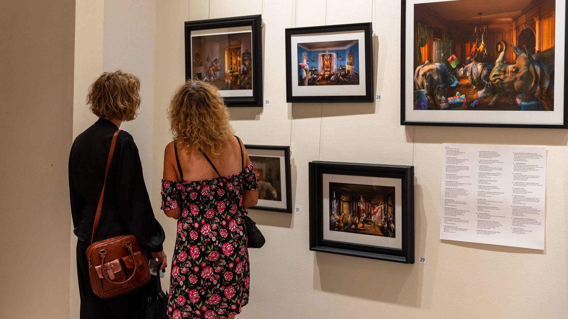 Two young people, both with medium length wavy blonde hair, looking at a gallery wall of framed photos. One is wearing a black jacket with a leather bag, the other is in a floral pink and black dress.