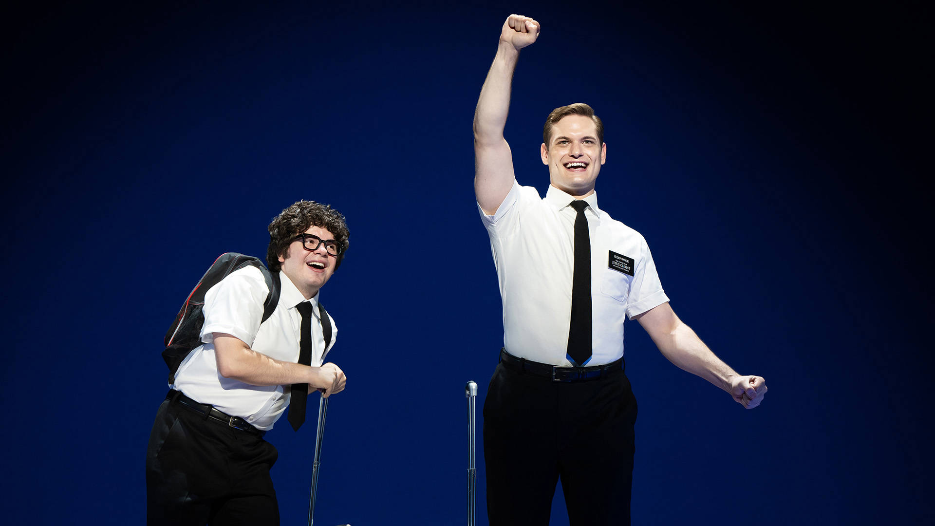 Two men with light complexion, one with dark curly hair and black-rimmed glasses, the other with short brown hair, stand together wearing white shortsleeve button-up shirts with black ties, suitcases in hand, exuding contrasting emotions—eagerness and wide-eyed anticipation. 
