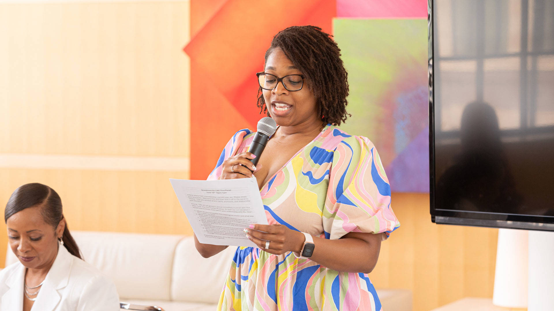 A young african american woman with medium length hair and a colorful top speaks into a microphone and is reading from a sheet of paper.