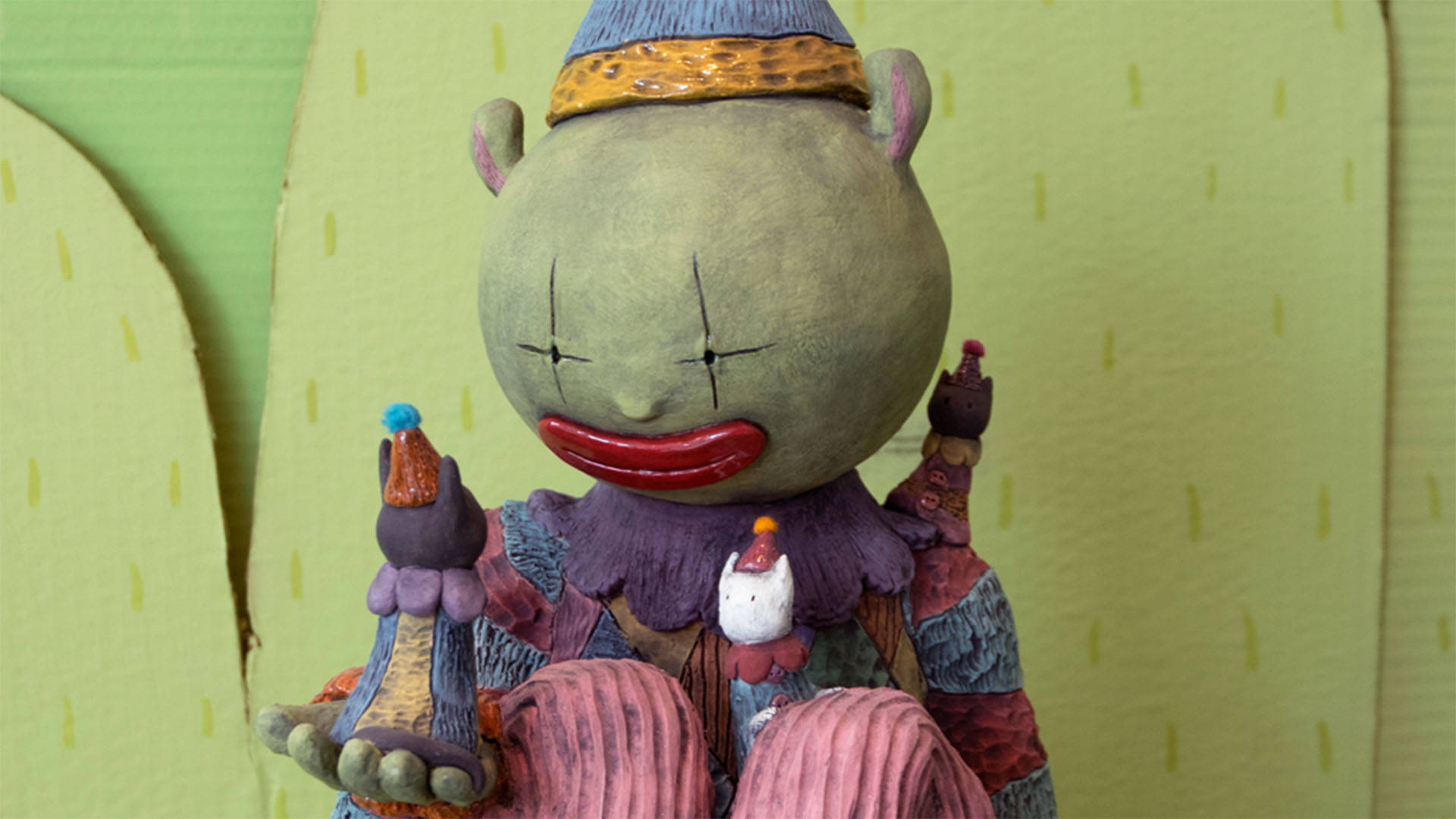 A ceramic clown sculpture with green skin sitting and holding a smaller cat clown in their hand.