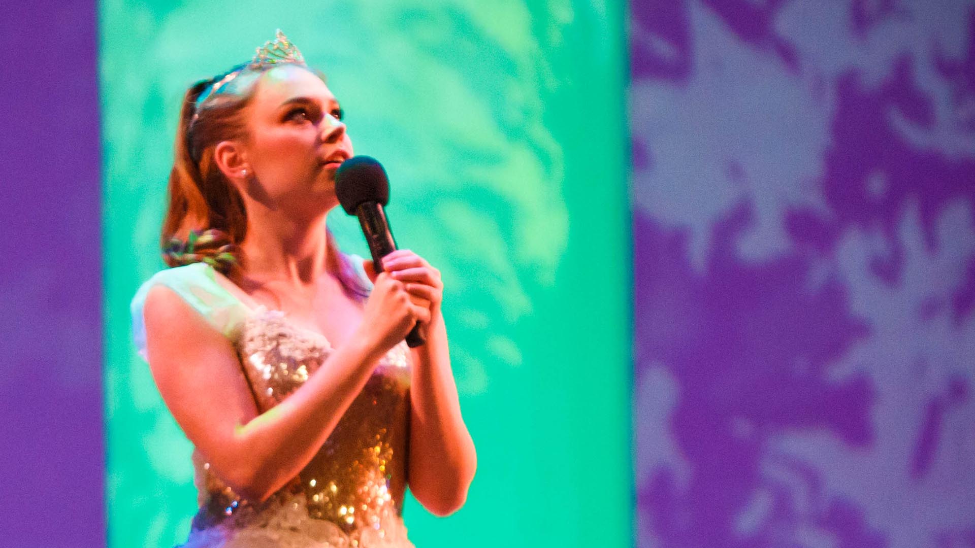 A young white woman with blonde hair in a gold sequin princess costume holding a microphone and looking up inquisitively. 