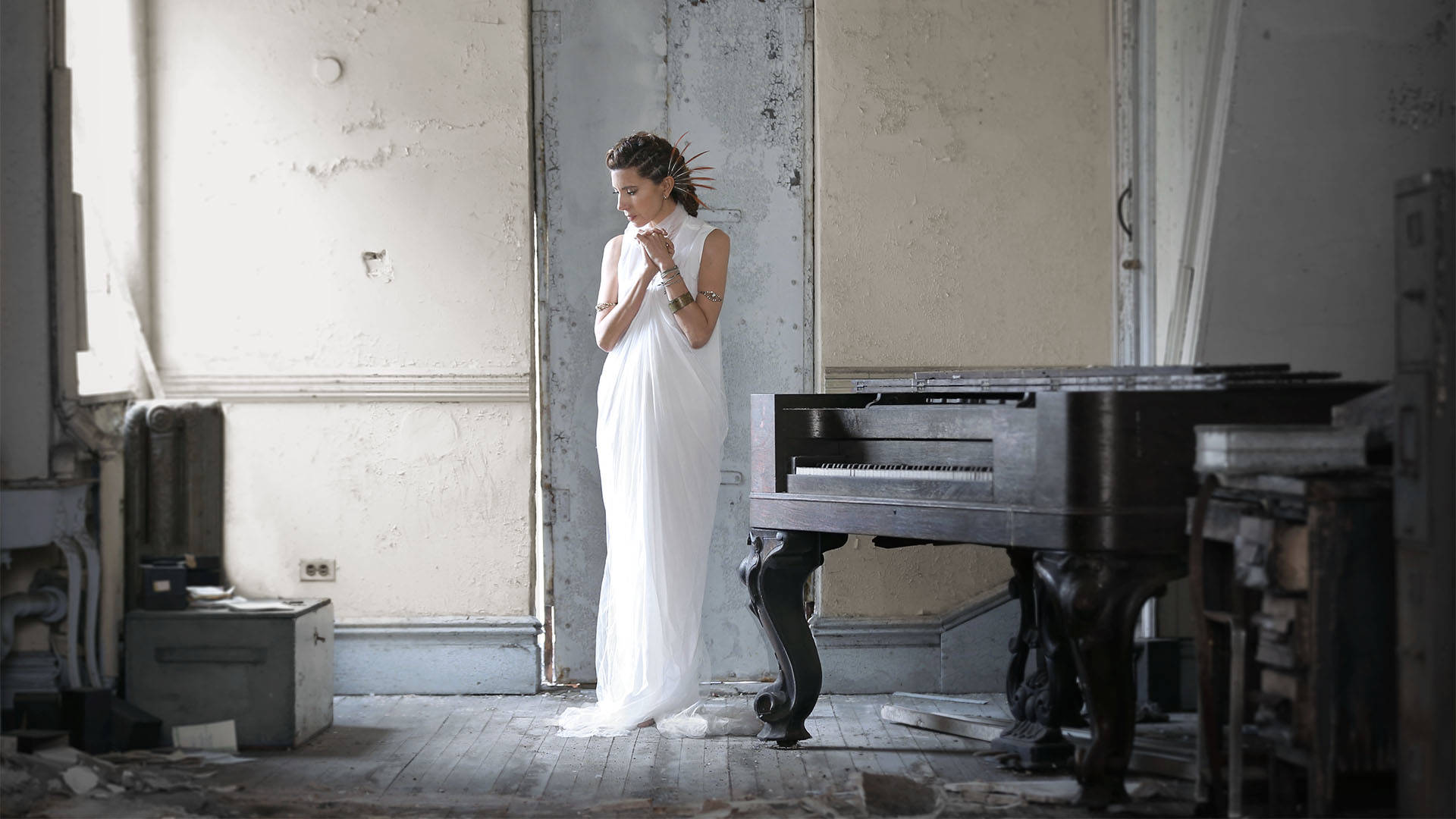 Magos Herrera, a Mexican woman with light brown long hair standing in a barren room next to a large piano. She is wearing a white sleeveless maxi dress and has many bracelets on her wrists along with feather pins in her hair.