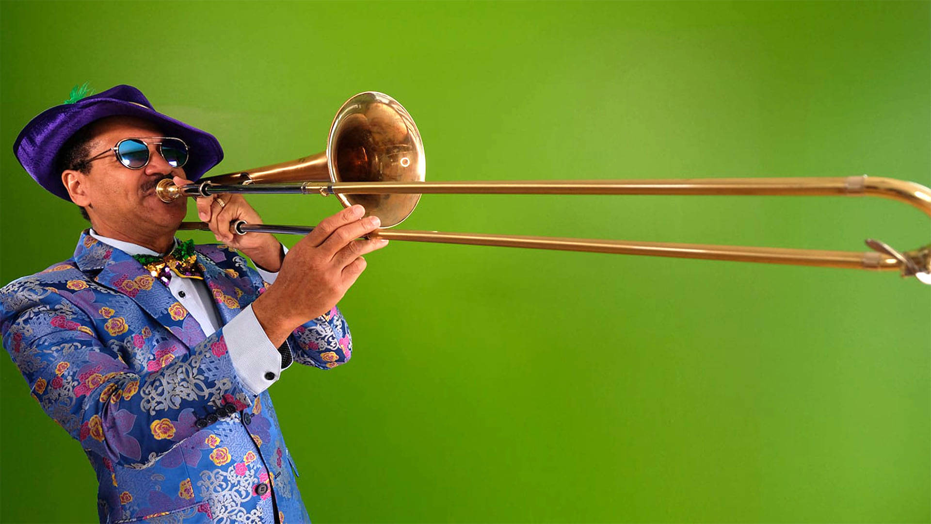 A Black man with short hair and sunglasses wearing a blue floral suit and purple festive hat, playing the trombone.