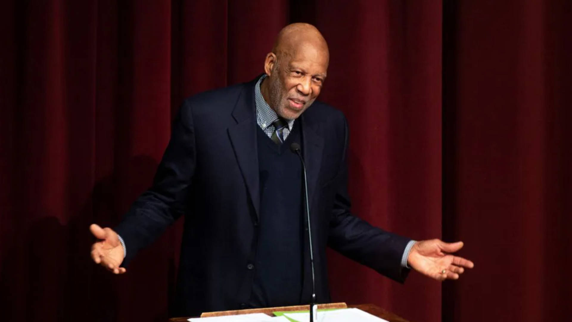An older african american man in a dark blue suit speaking at a podium in front of a red curtain.