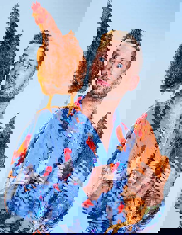 Headshot of a young caucasian man with short blond hair and a blue robe with chickens designs. He is holding a hen and another is standing on his shoulder. The background is blue.