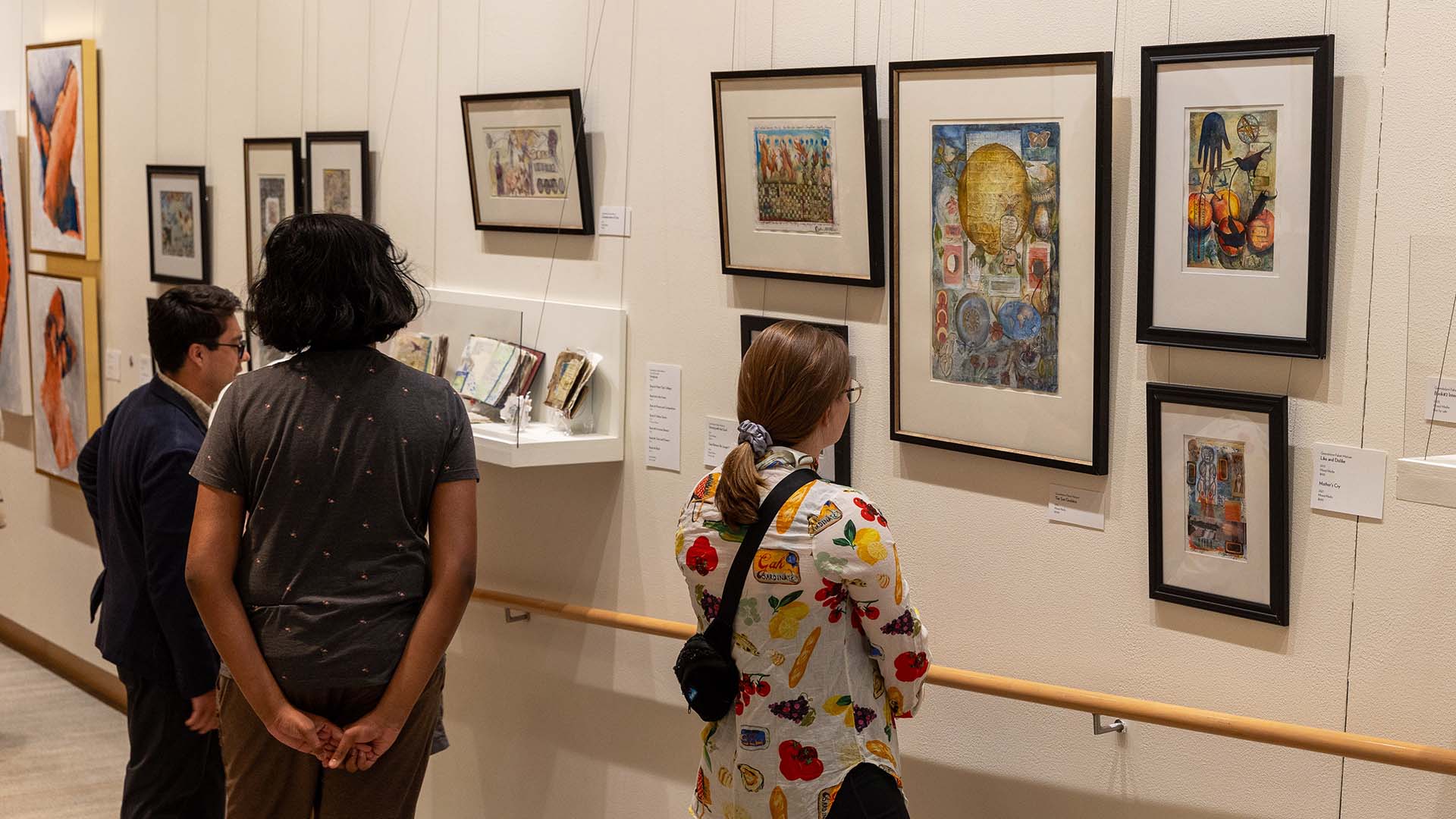 Three people standing in a hallway gallery with white walls, displaying a collage of framed art on the wall.