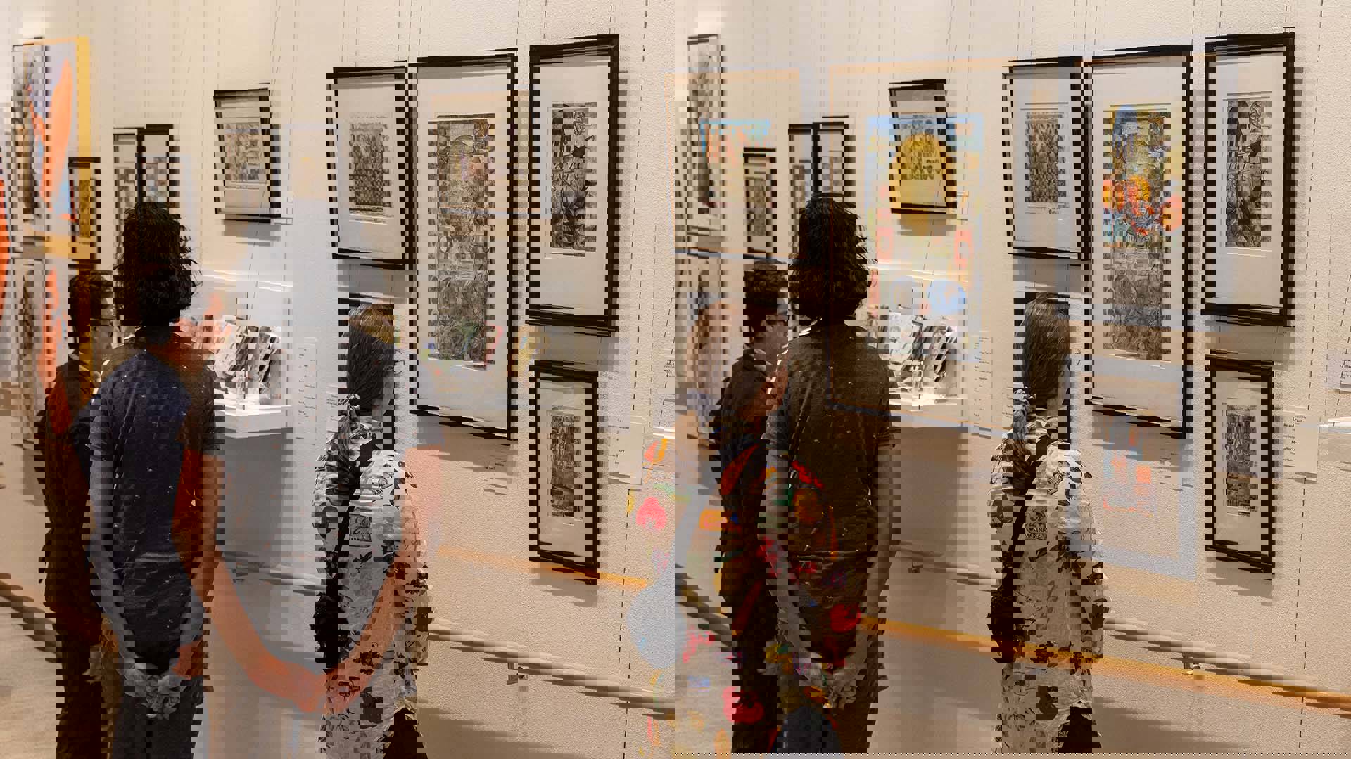 Three people standing in a hallway gallery with white walls, displaying a collage of framed art on the wall.
