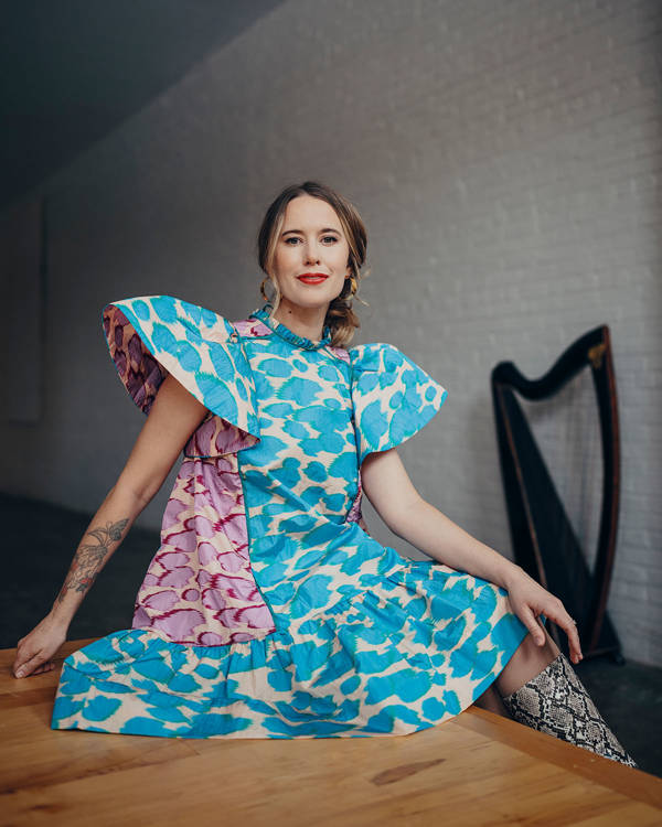 A caucasian woman with dark hair with blonde highlights and brown eyes is smiling and looking toward the viewer. She is wearing a dress with large shoulder cuffs, and blue and purple splotches. A harps is in the background.