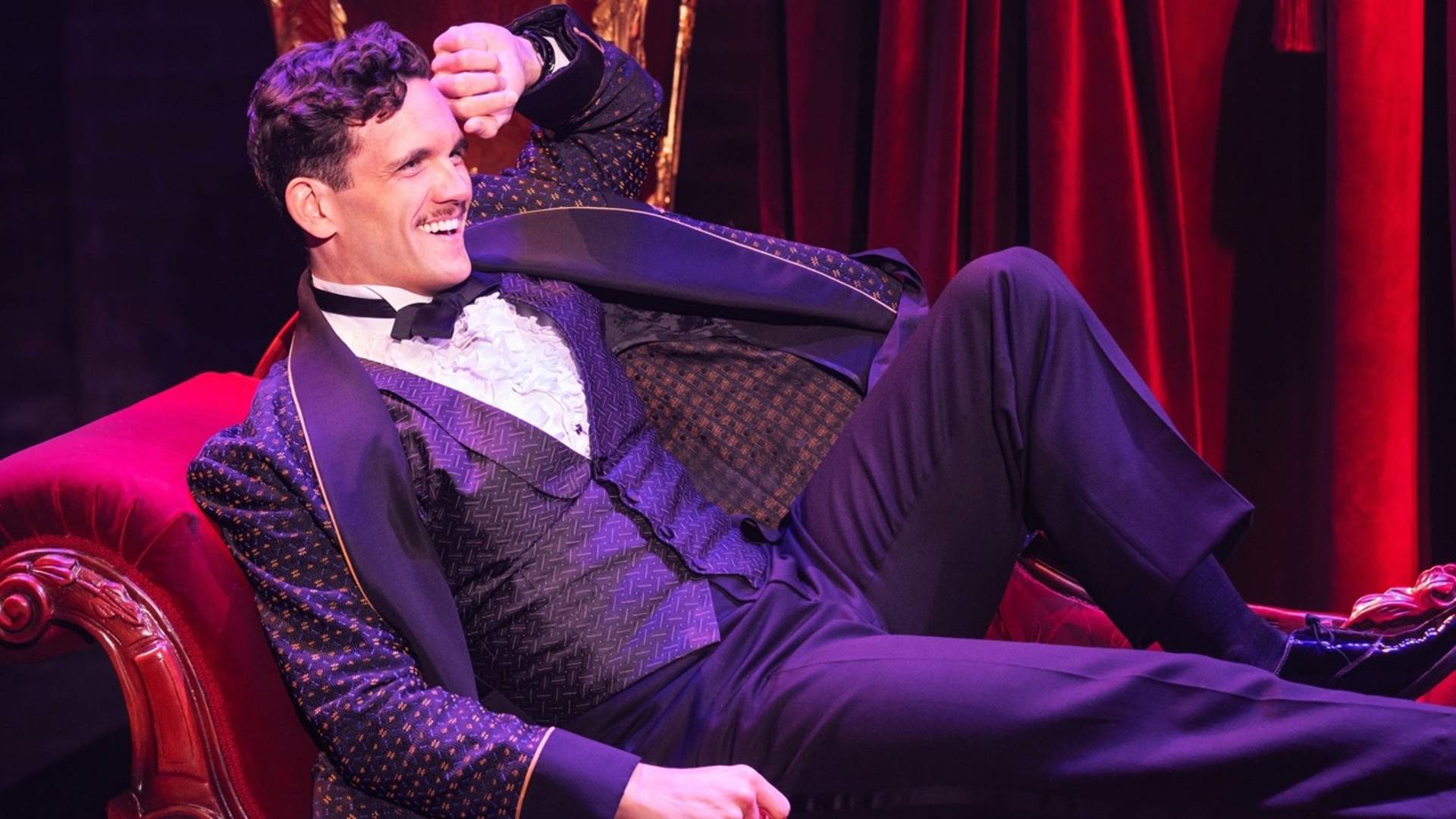 An actor in Funny Girl seated on a red velvet chaise lounge in a fancy tuxedo smiling with his knee up. He is of light complexion, with short brown hair and a moustache.