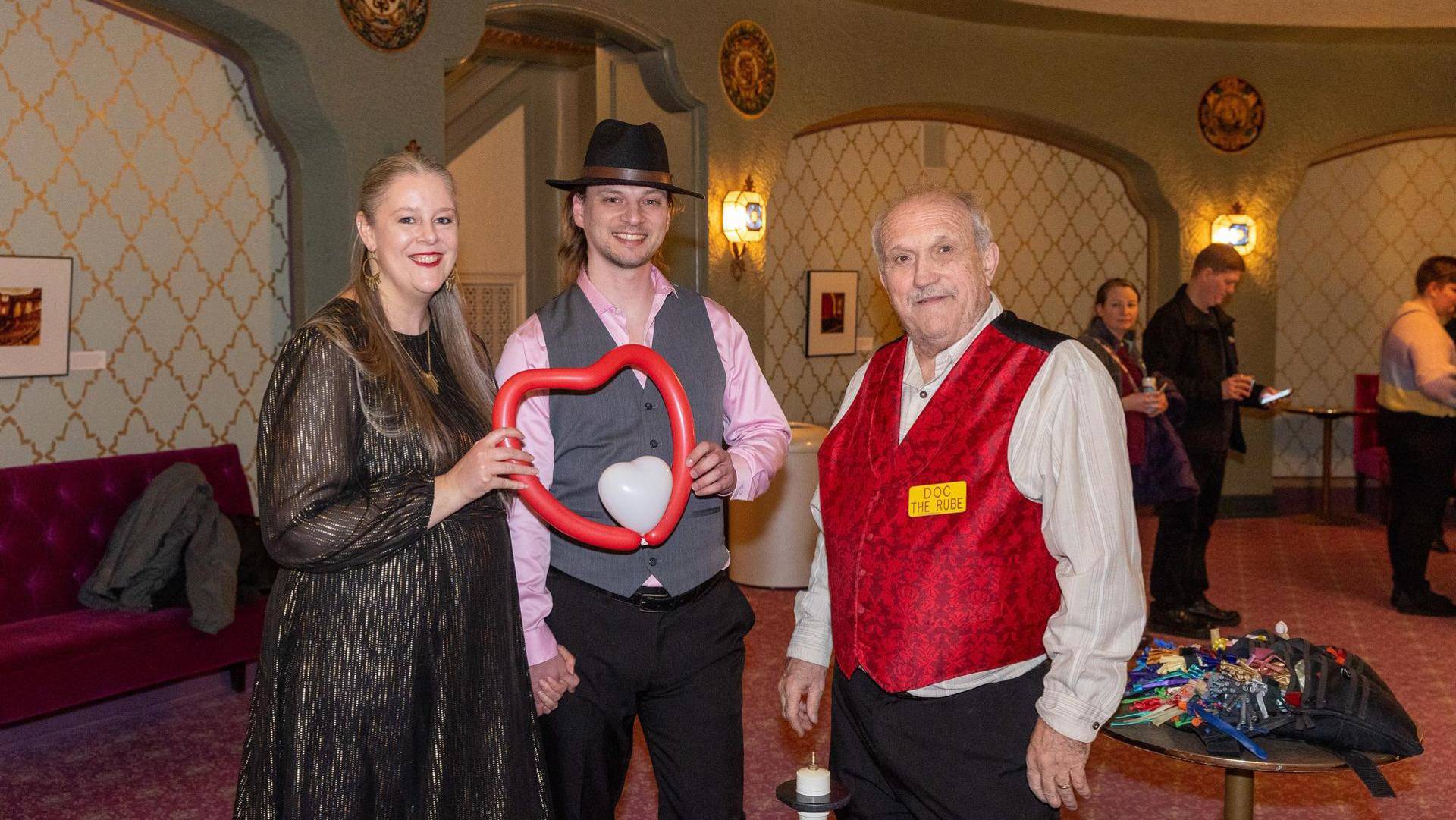 A man and a woman stand next to an older balloon artist in a red vest. The couple is holding a heartshaped balloon.