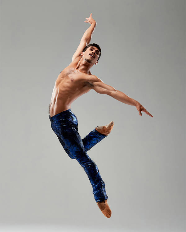 A fit man with medium complexion and short dark hair striking a pose midair against a grey background. He is wearing blue pants and is shirtless.
