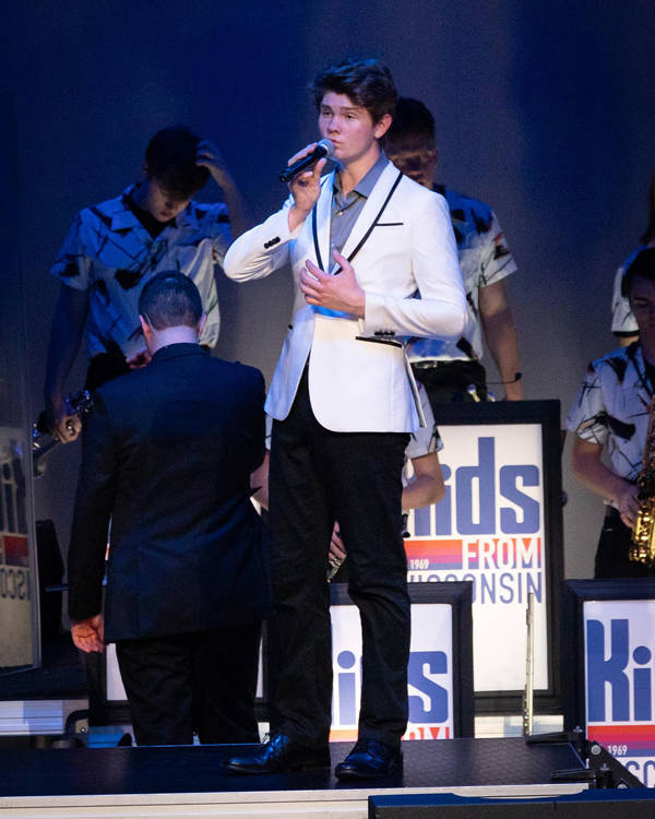 A performer with light complexion and dark hair singing into a microphone with a white tuxedo on.