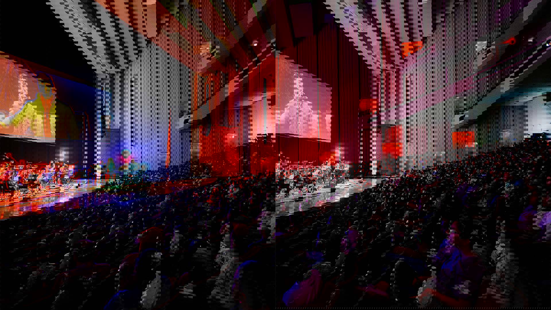 A full house audience watching an orchestra performing on stage under a large screen with a scene from Spiderman into the Spider-verse.
