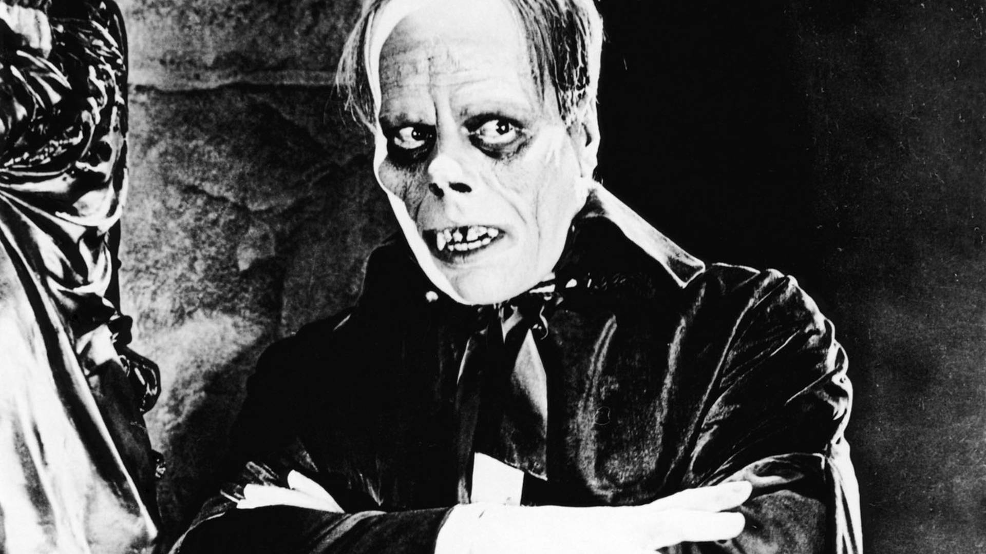 The phantom of the opera has scary looking face with fangs and dark eyes, wearing a black cloak and crossing his arms.