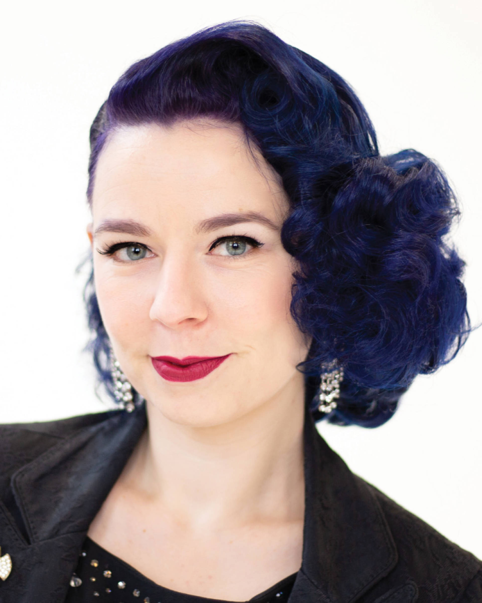 Headshot of a caucasian woman with dark blue hair, blue eyes and a dark top and jacket.
