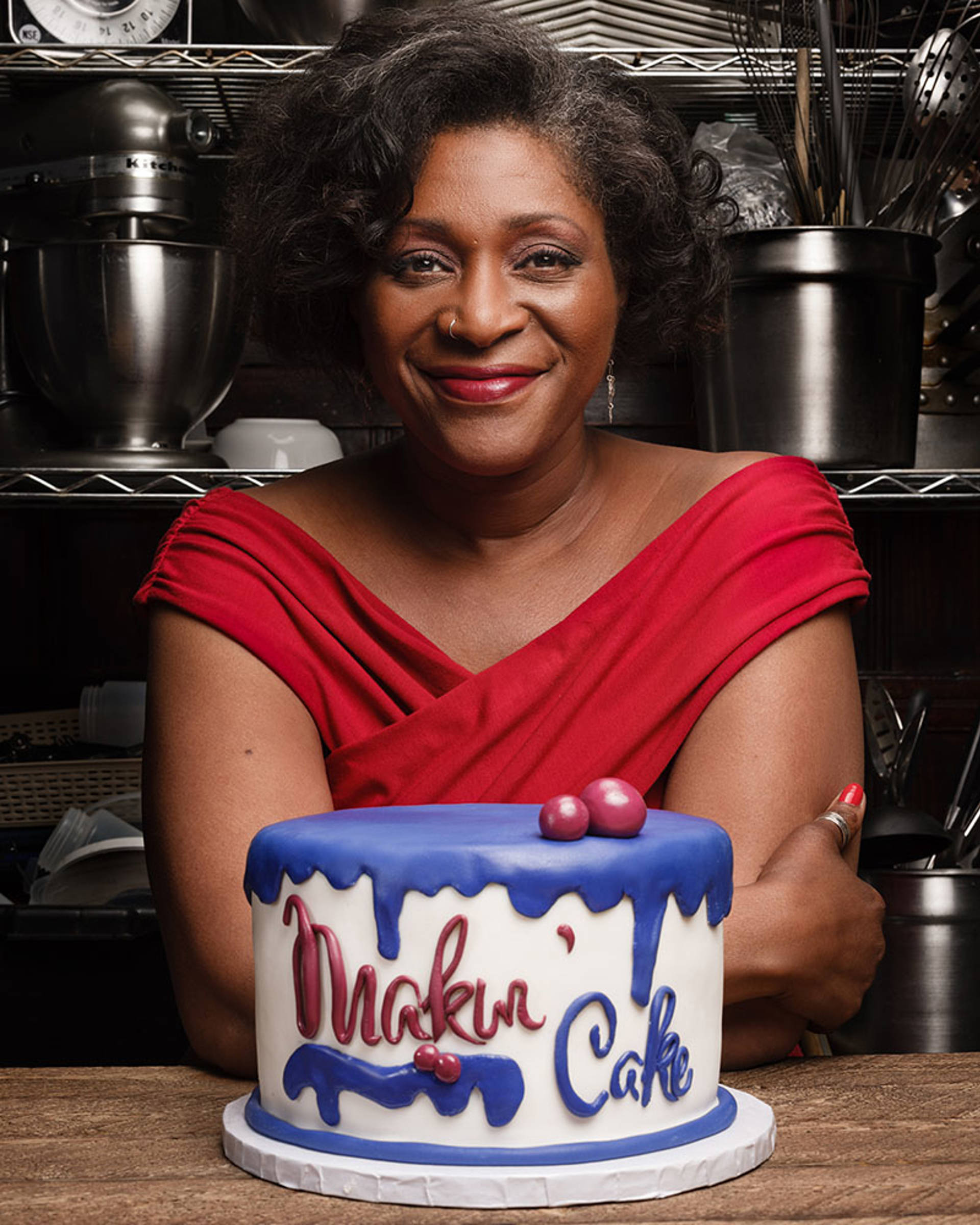 An African American in a red dress with medium length hair smiling as she sits behind a cake decorated with 'Makin' Cake' frosting text