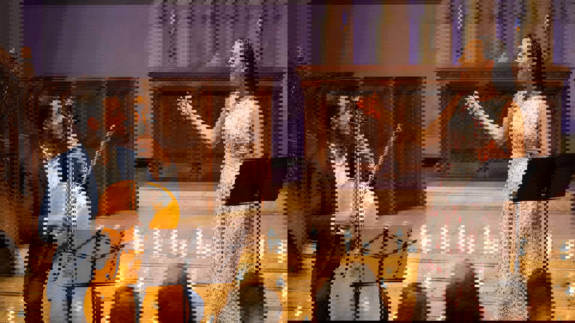 A man with light complexion, dark hair and a light beard in a blue suit playing a cello. To his left is a young woman with light complexion, long brown hair, and a white dress. She is singing.