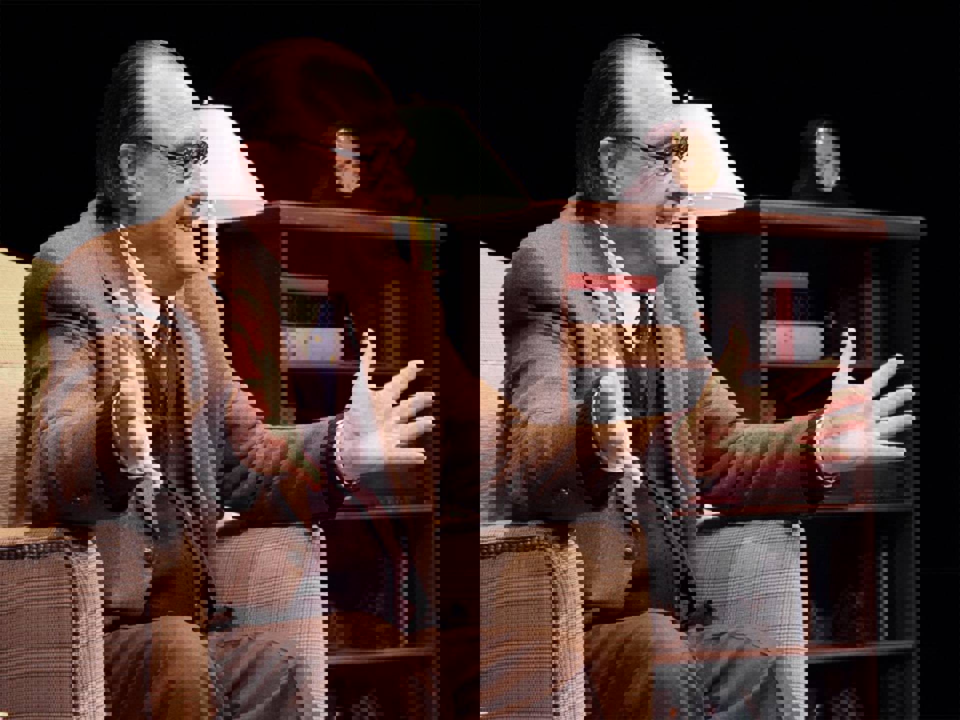 An older, white man with a receding hairline and black glasses in a tan suit. He is sitting in a plaid chair with his hands held out in front of him. Behind him is a lamp and bookshelf.