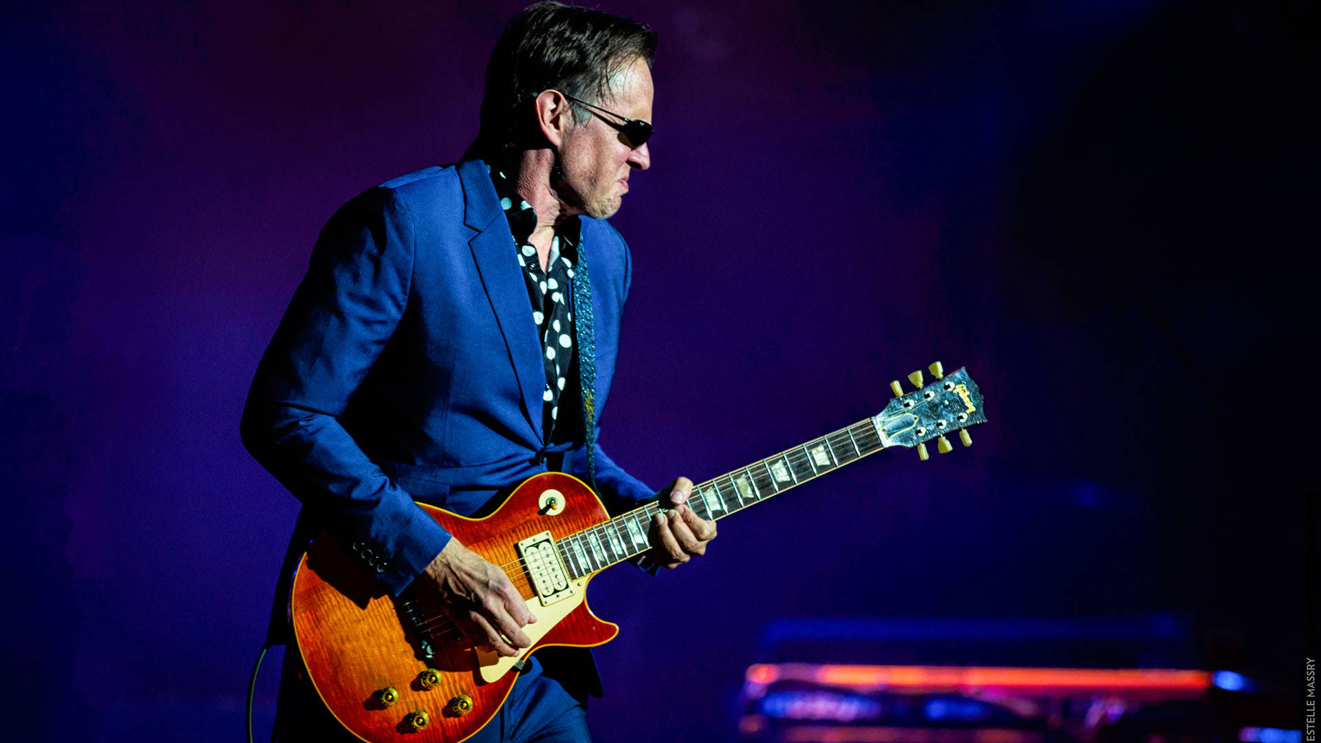 A caucasian middle aged man performing on stage in a blue suit, black and white polka-dotted shirt, playing an orange electric guitar.
