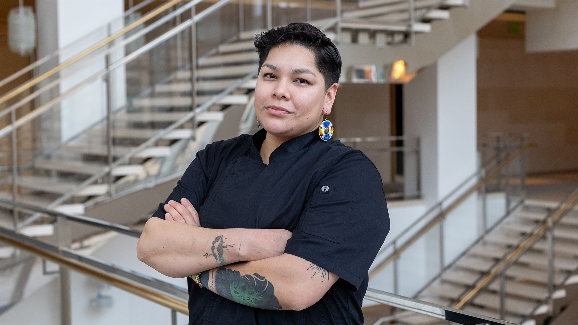 A medium complexion person with short black hair wearing a black chef coat and dangling blue and orange patterned circular earrings. Her arms are crossed, showing a few tattoos, in Overture Hall Lobby.