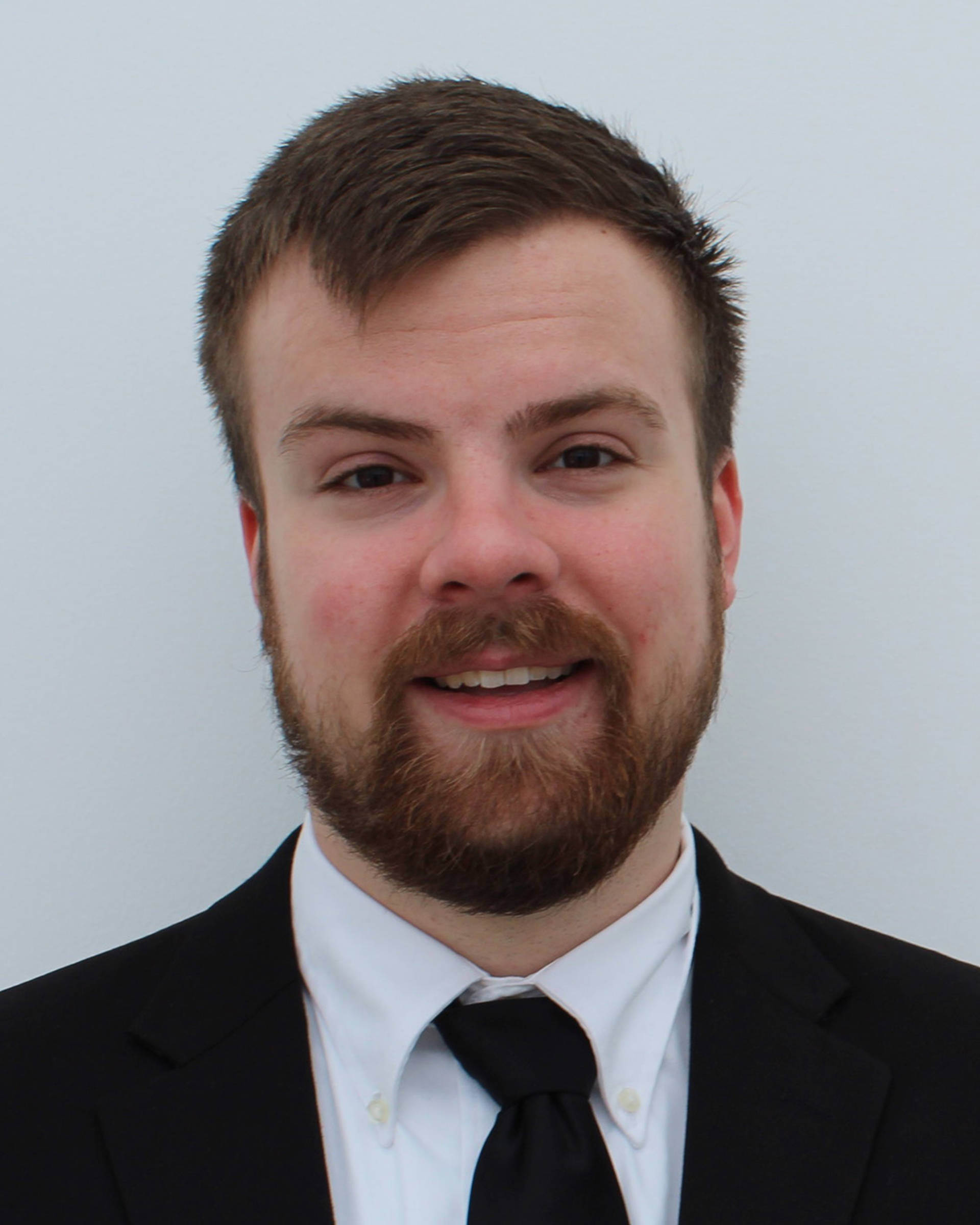 Headshot of a caucasian man with short brown hair, brown eyes, a beard, and a suit with black tie.
