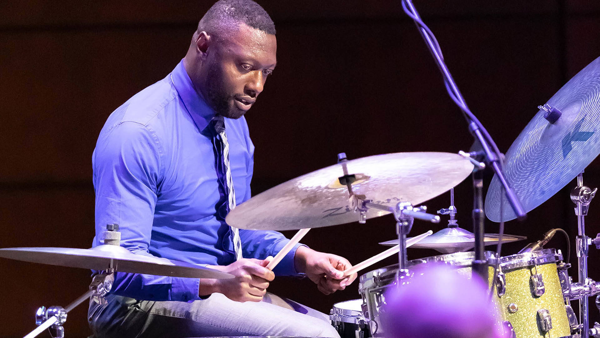 A Black man in a blue shirt and tie playing a drum set.