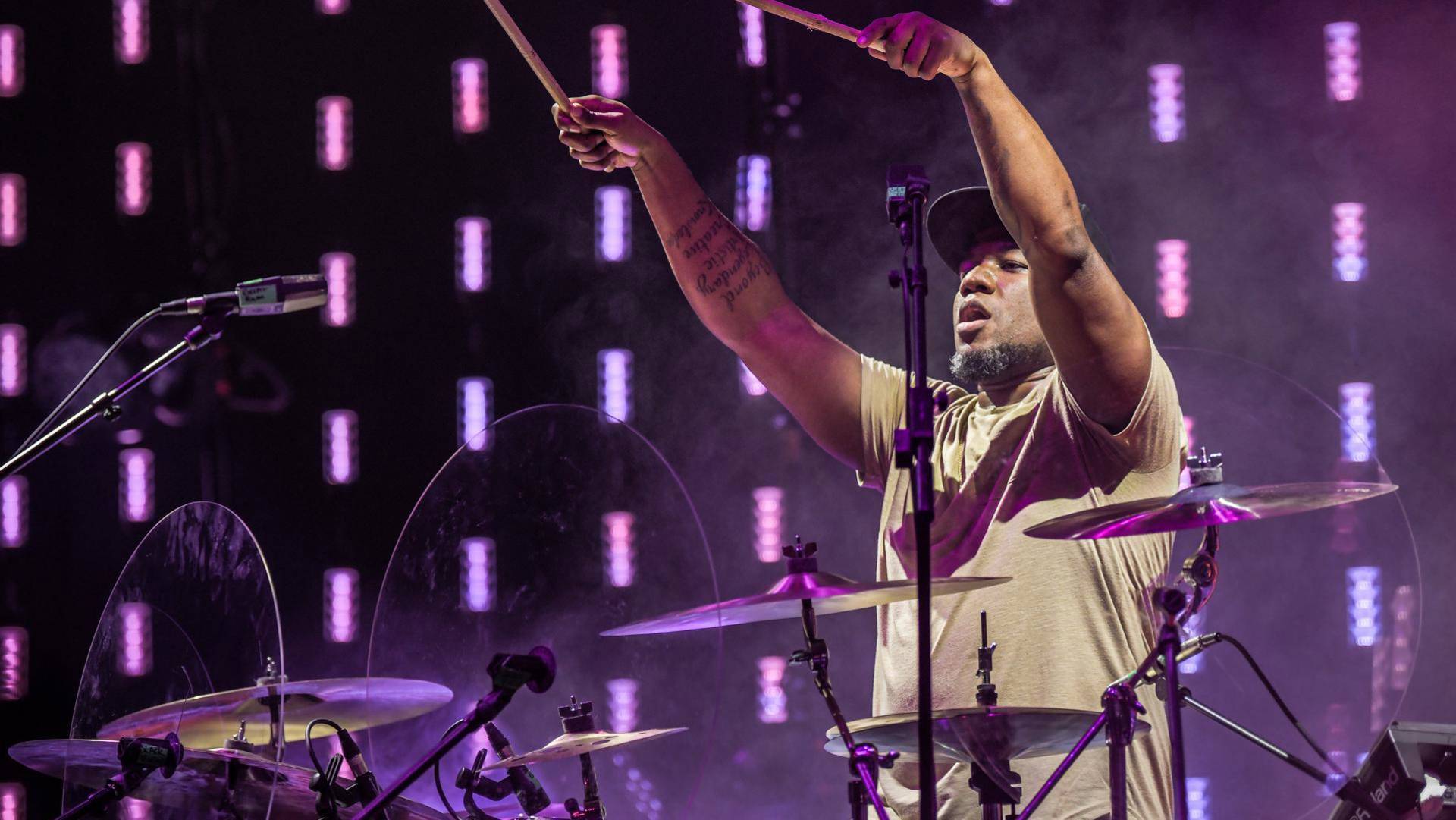 A drummer standing behin his drum set and pointing his sticks outward. He has dark complexion, a tan shirt, a short beard and a dark baseball cap.