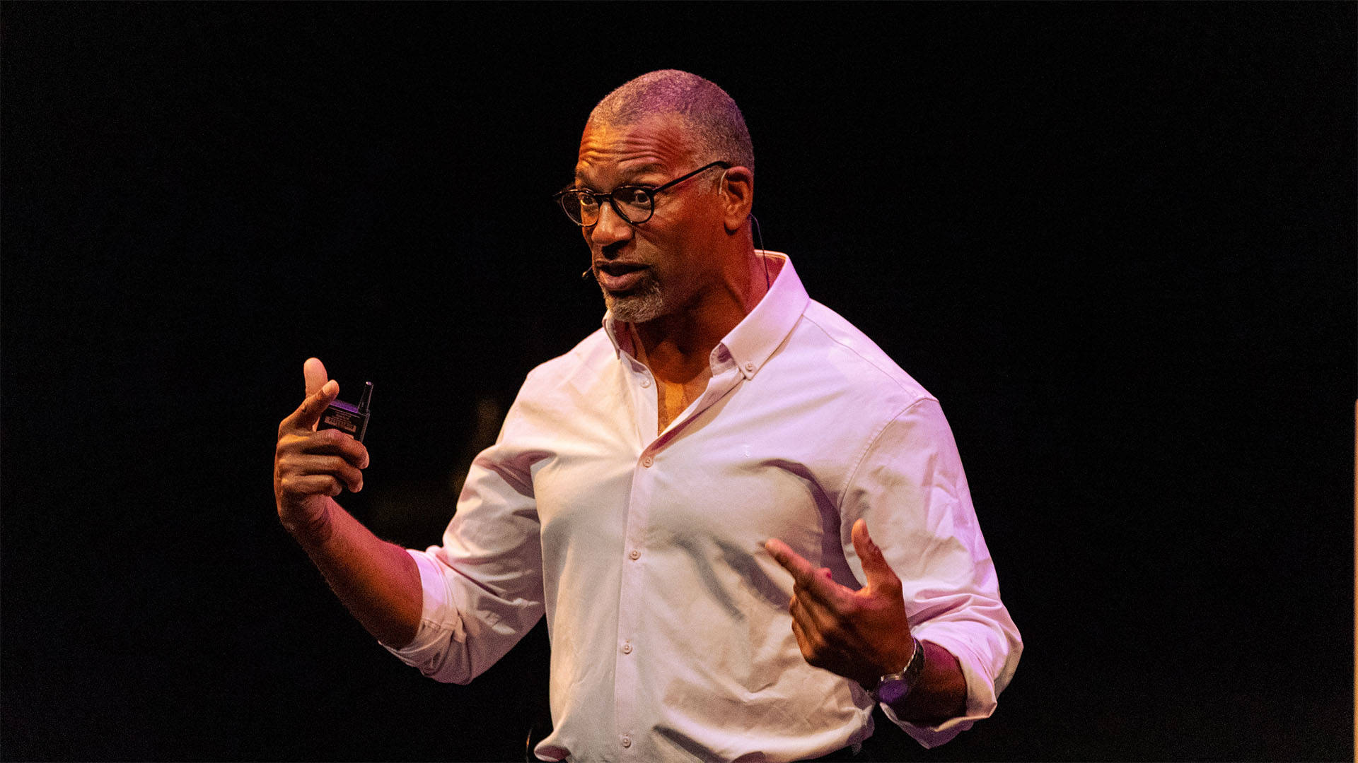 An african american man with buzzed greyish hair, glasses, and a white button-up shirt speaking against a dark background