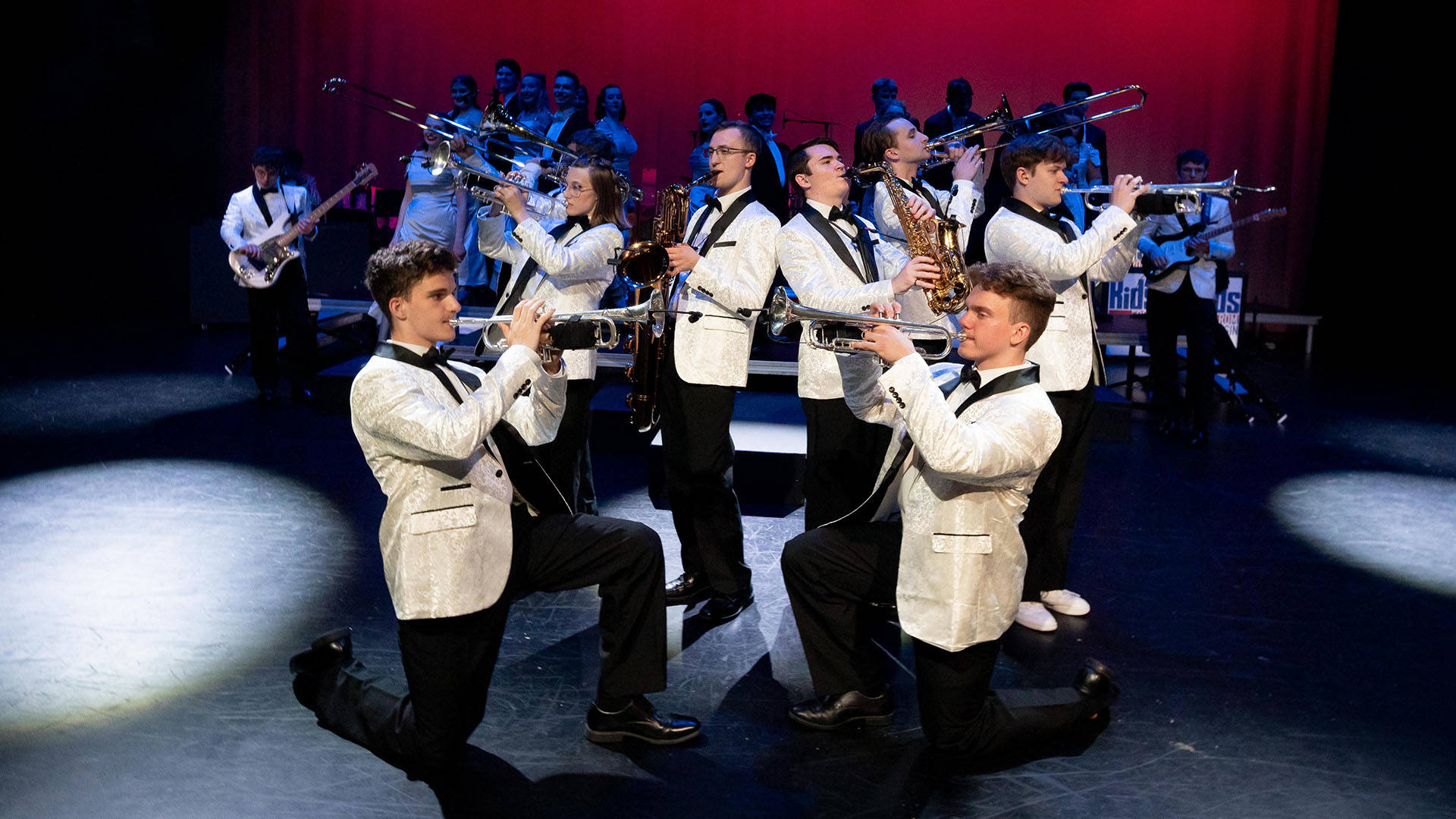 The horn section of Kids from Wisconsin performing on stage in white suits with black slacks.
