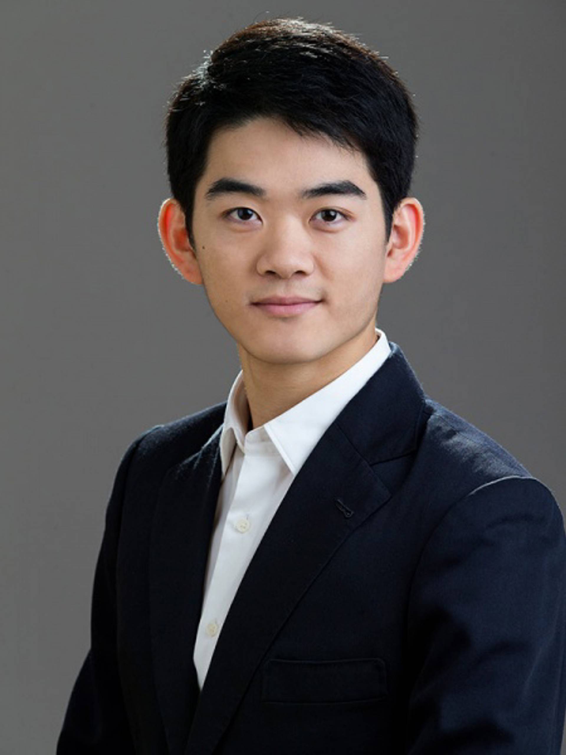 Headshot of a young man with medium complexion, short black hair, a white collared shirt and a dark jacket against a grey background