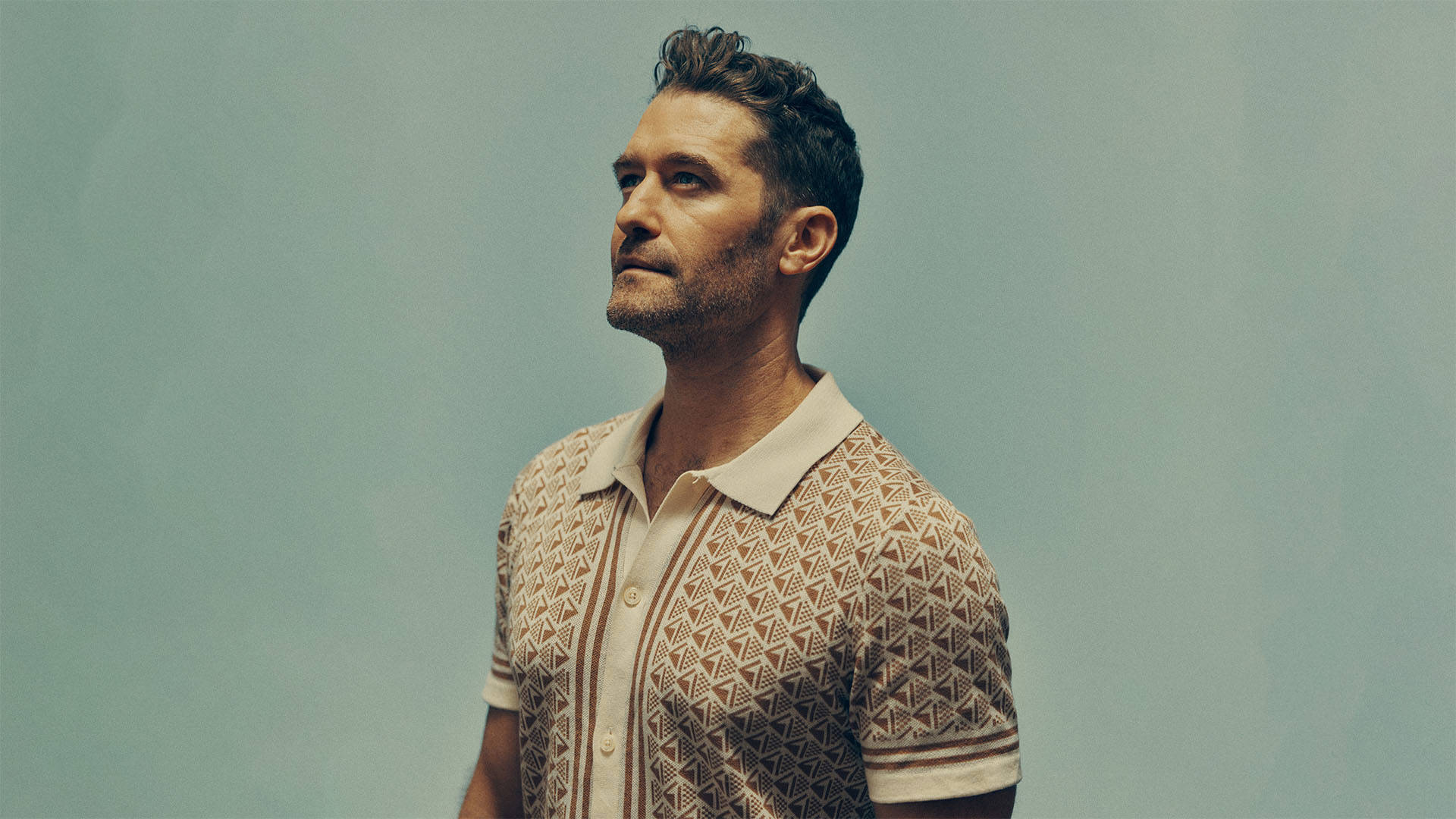 Matthew Morrison, a caucasian man with short brown hair and trimmed facial hair, wearing a tan and cream button up knit polo, looking slightly upwards. A grayish teal backdrop is the background.