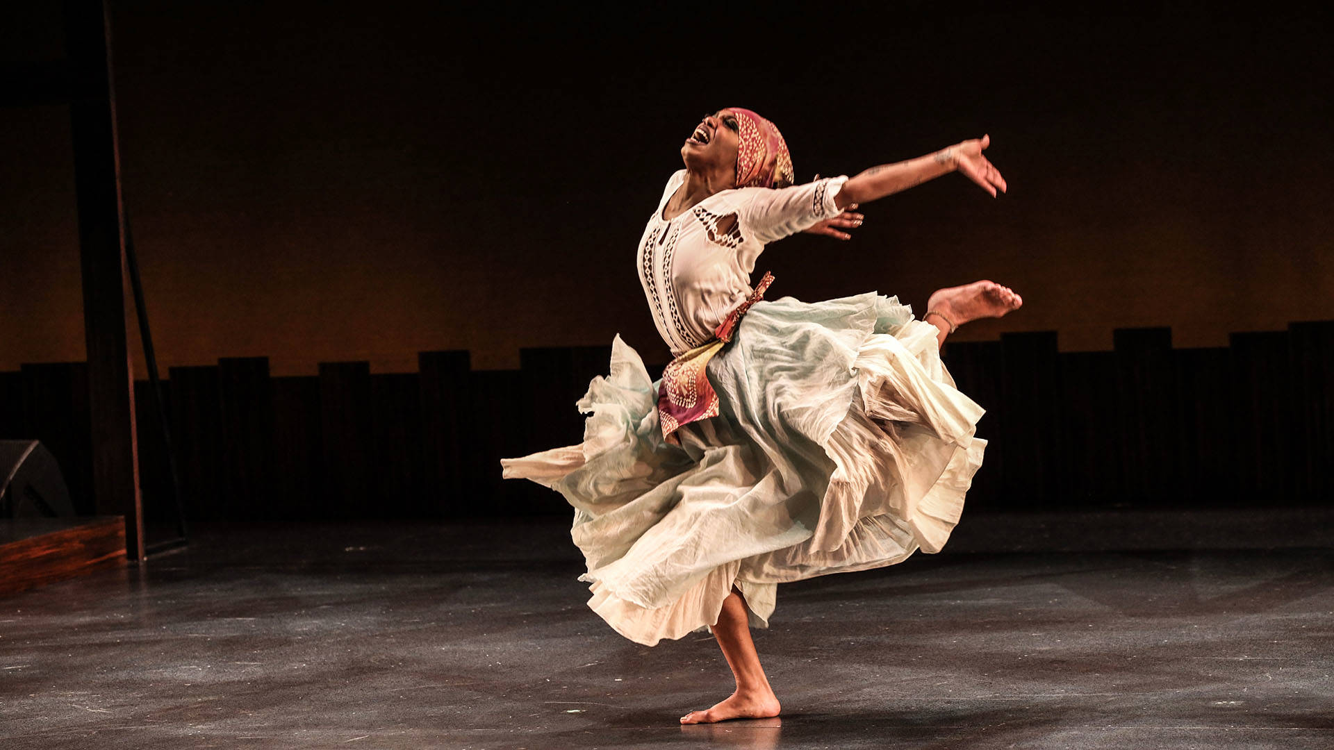 A Step Afrika! dancer performing on a dark stage. They have dark complexion, a flowing beige dress, and reddish headwear. Their arms and left leg are outstretched as they dance.