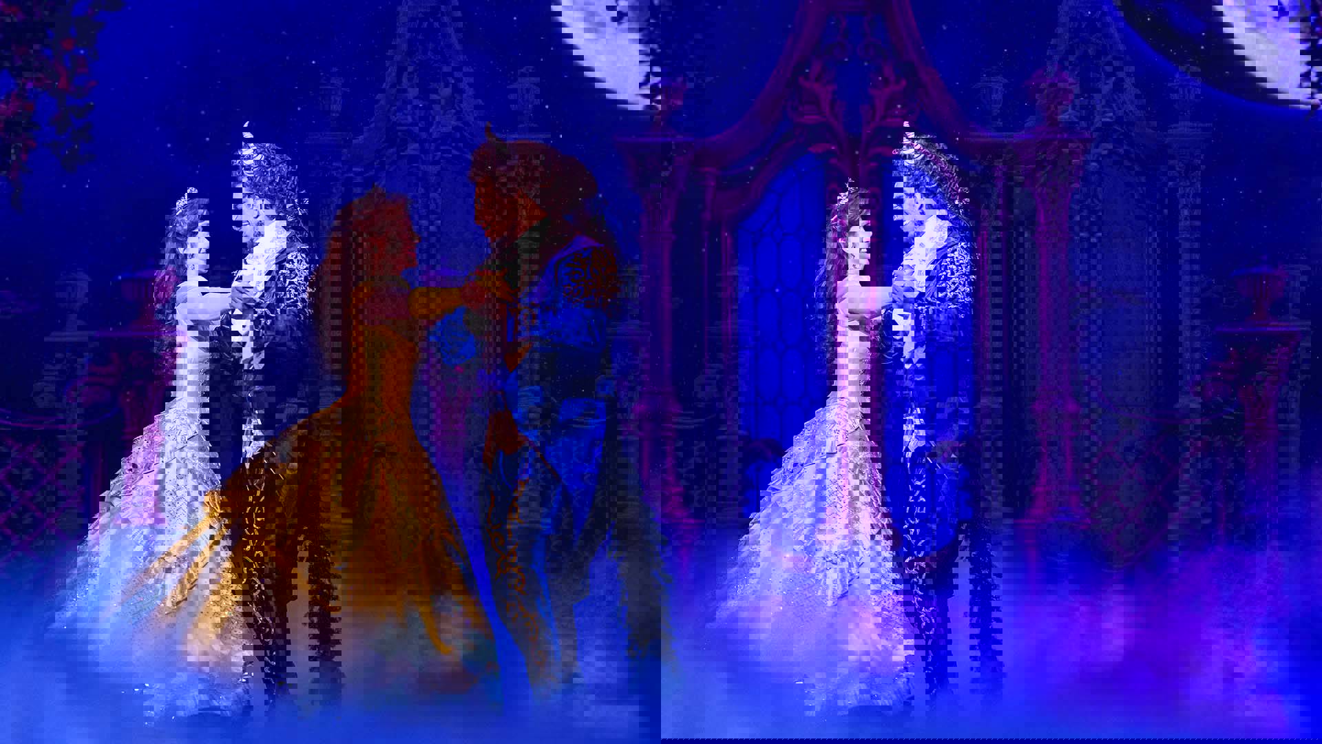 A woman with light complexion and brown hair wearing a yellow ballgown, and a beast-like man with long hair and horns in a blue and yellow suit, dance in front of glass double doors.