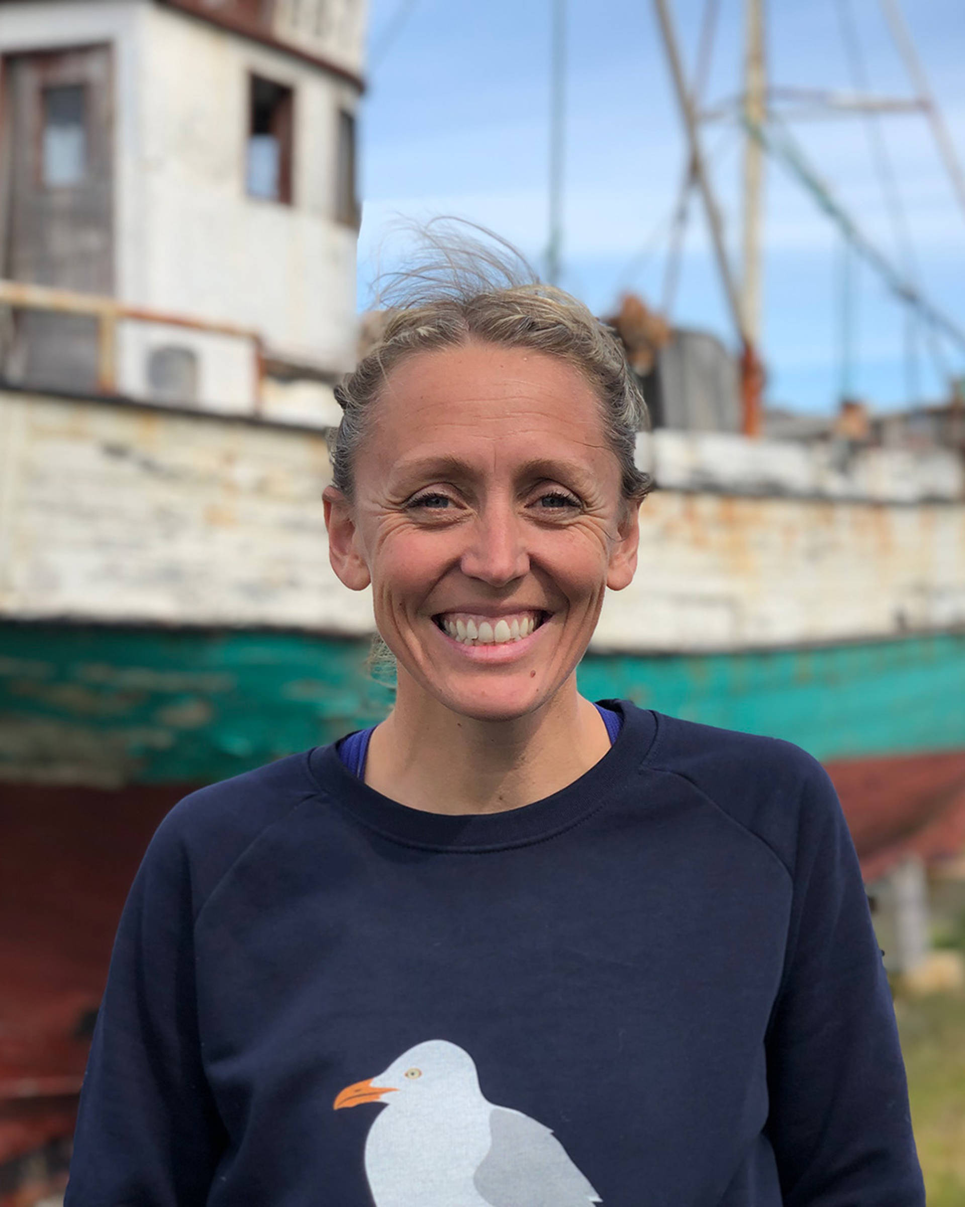 Headshot of Lucy Hawkes. She is smiling warmly toward the camera, has pulled-back hair, and a blue sweater on with a seagull graphic on it