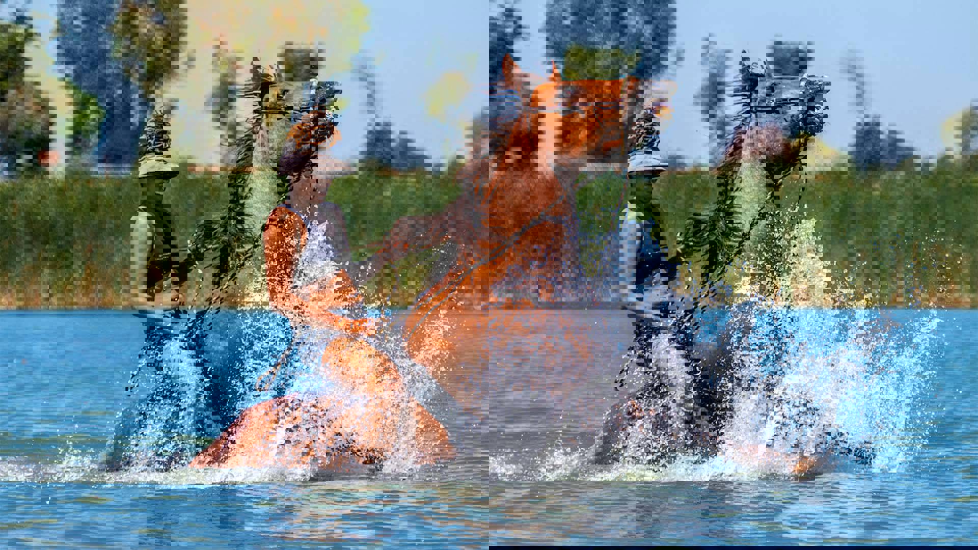 A medium-skinned person with brown hair in a bun and visor riding a brown horse in water.