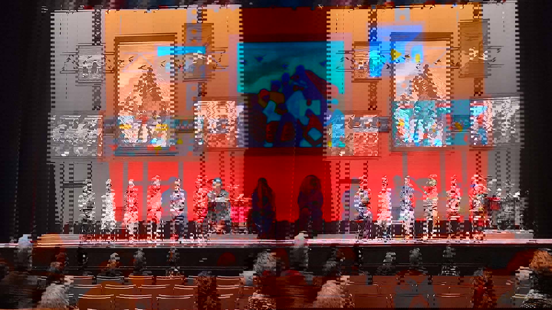 A group of Step Afrika! dancers sit on a stage and talk to audience members with vibrant African art as a backdrop.