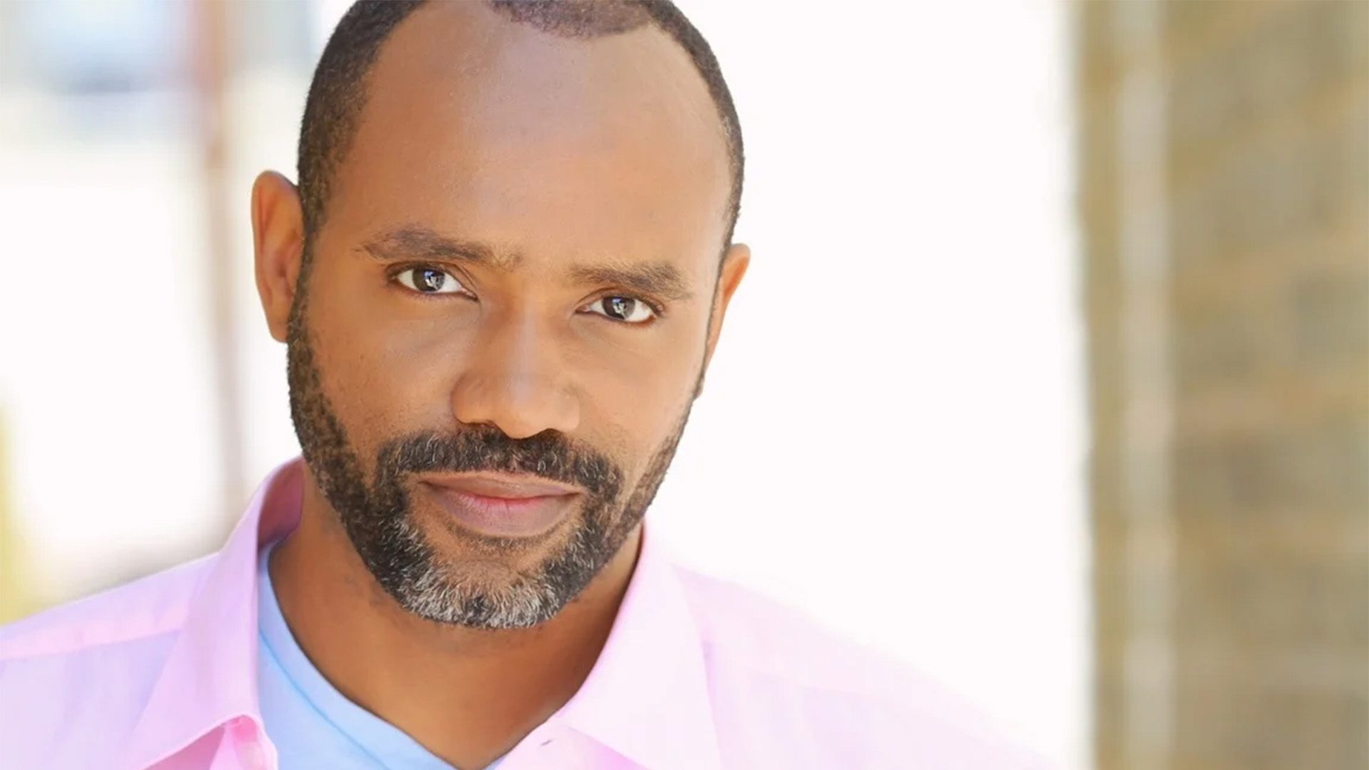 A headshot of a Black man with dark, buzzed hair and facial hair. He is wearing a pink, button-down shirt over a light blue undershirt.