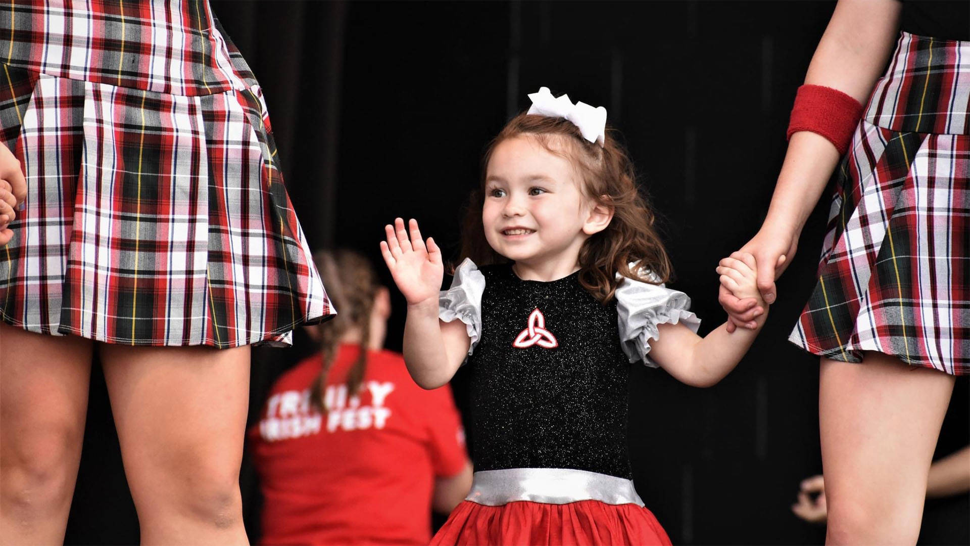 A young girl in a black, red and silver costume smiles and waves while holding hands with two older performers in plaid skirts during a dance performance.