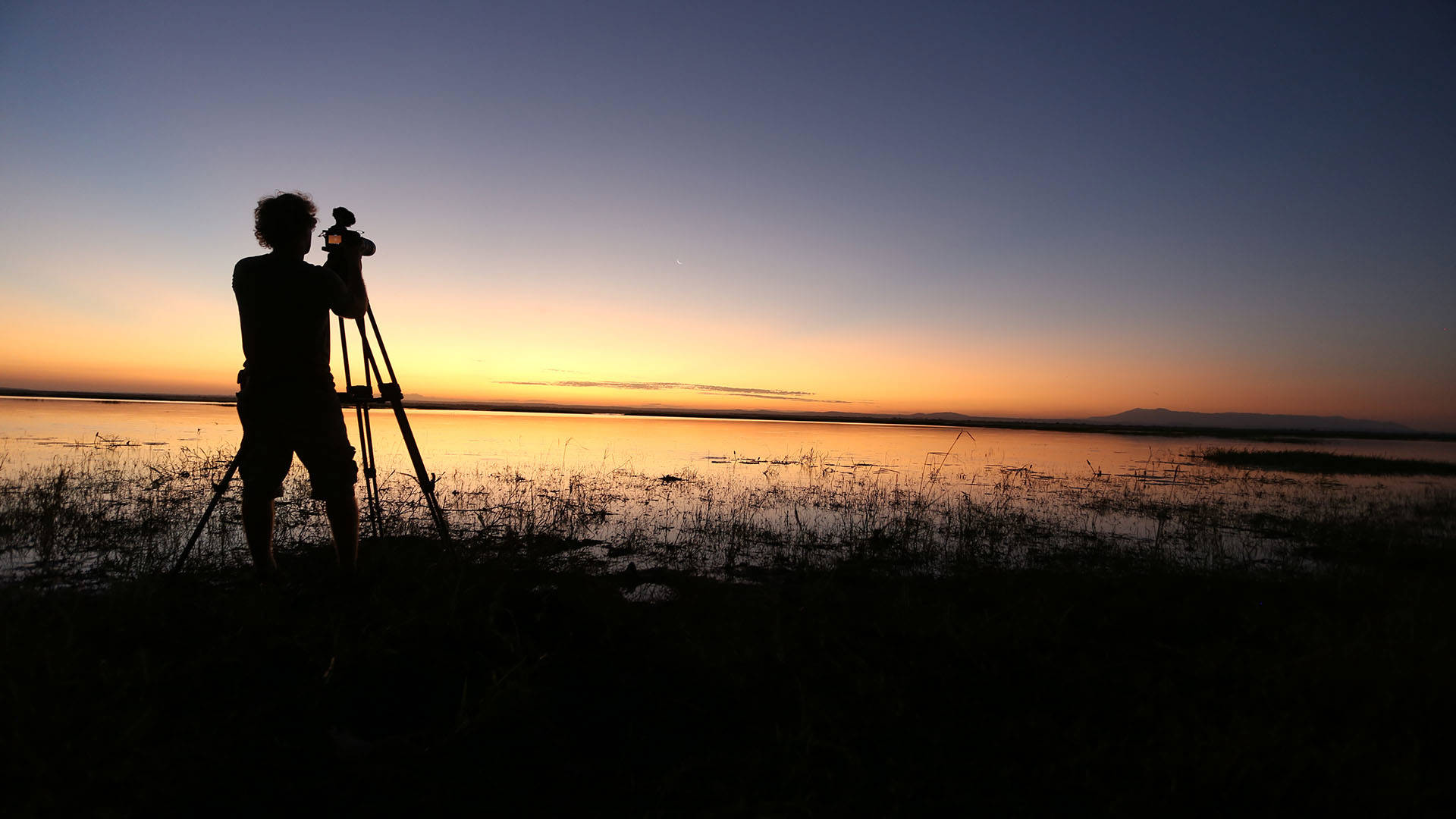 silhouette of a videographer with a camera at dusk next to a body of water.