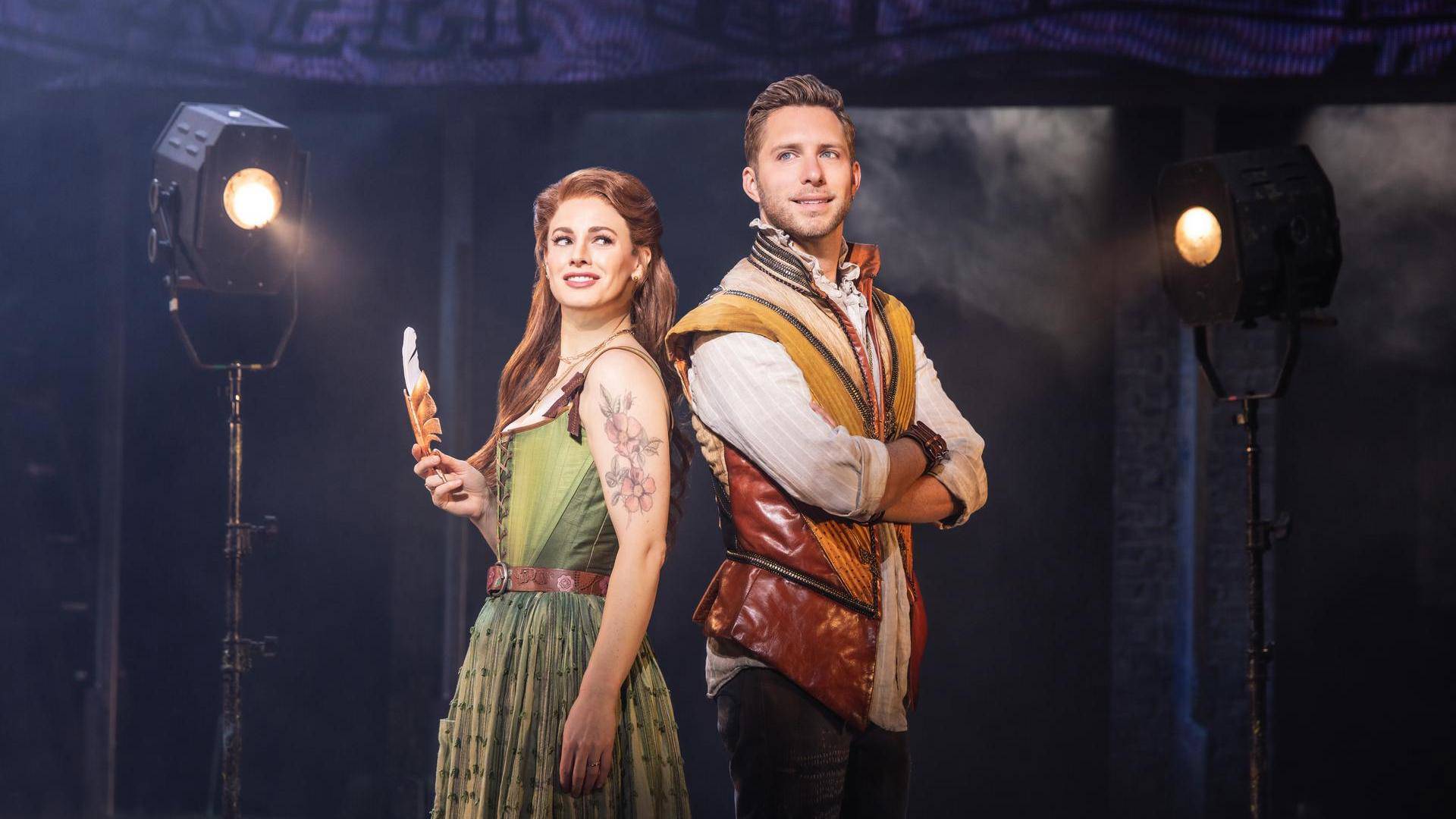 A woman with light complexion, reddish hair and a green dress stands back to back with a man in a yellow and brown vest with light complexion and short brown hair crossing his arms. The woman is holding a quill.