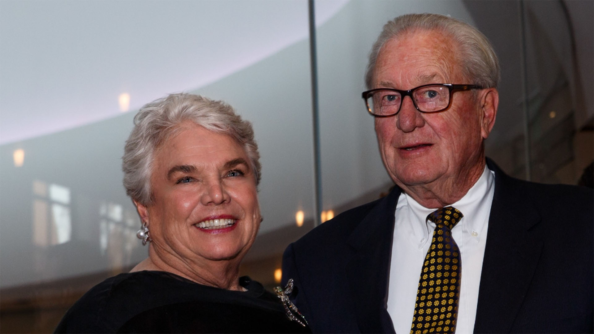 An older caucasian woman and man, both with short white hair, wearing formal clothing and smiling for the camera.