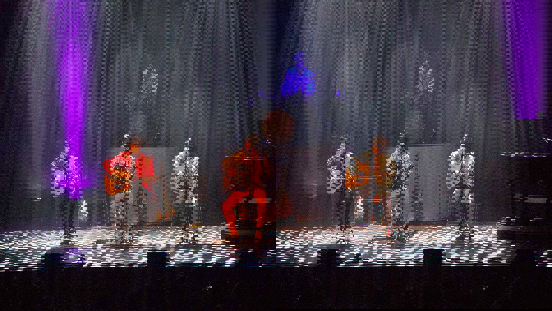 Beatles impersonators wearing colorful clothing and sitting with acoustic guitars on a stage.