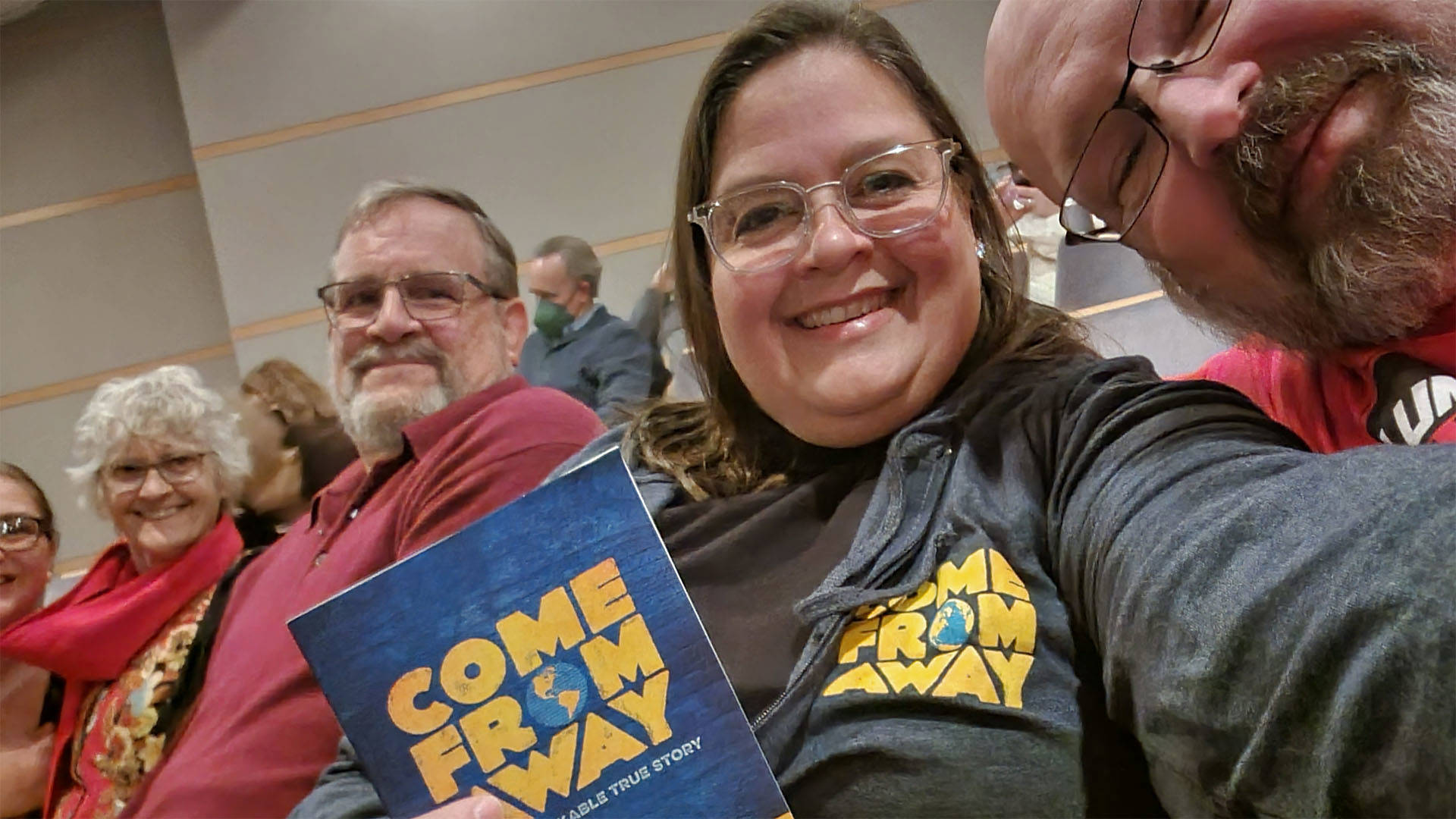 A woman with light complexion, longbrown hair and glasses sits in a theater and smiles towards the viewer. She is wearing a hoodie branded with the "Come From Away" logo and holding a program for the same show. Other seated audience members in her row are smiling towards the viewer as well.