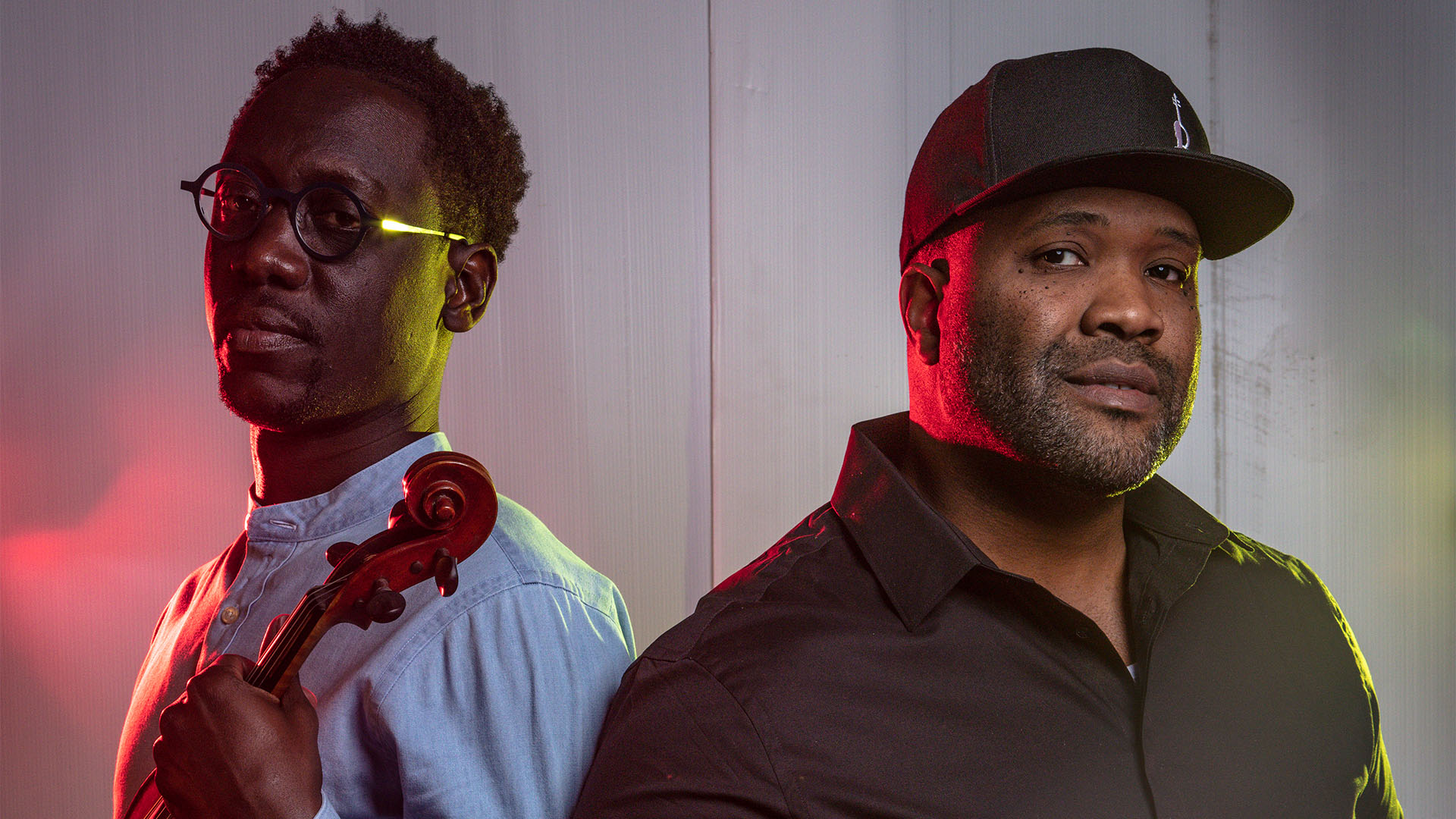 A posed shot of the two lead members of Black Violin. On the left, a man with dark complexion, glasses, short dark hair and a light shirt. He is holding a violin. On the right, a man with dark complexion, a dark baseball cap, and a dark collared shirt.