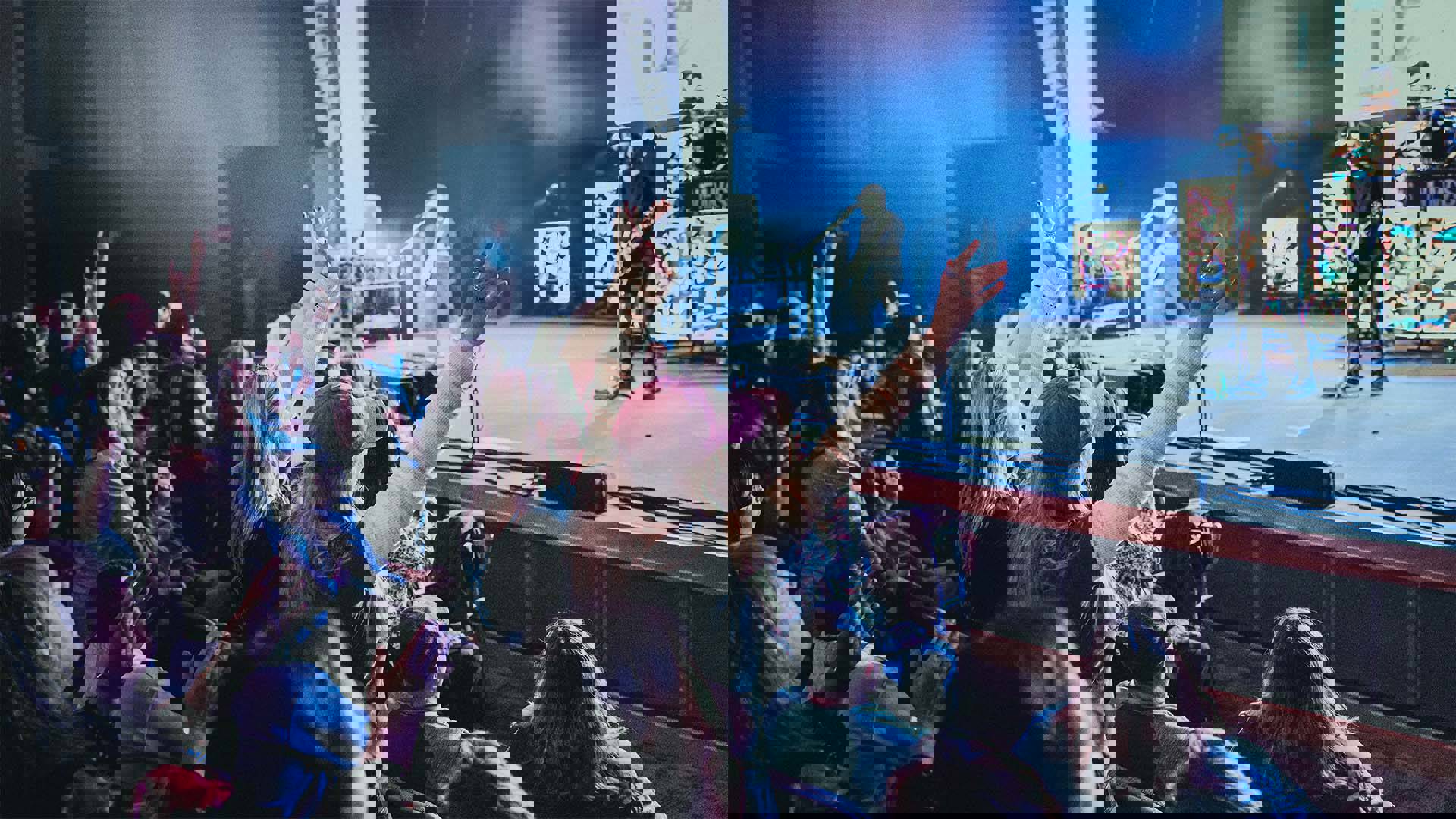 An audience watching a rock band on stage. Several have their hands in the air.