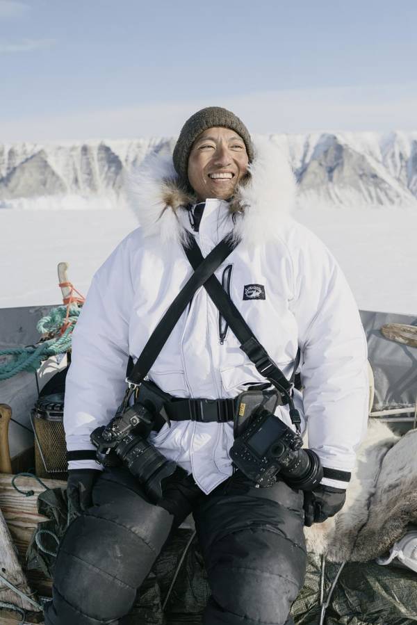 Photographer Kiliii Yuyan smiles while sitting with gear in front of an arctic mountain scape. He wears a white coat, tan hat and has two cameras.