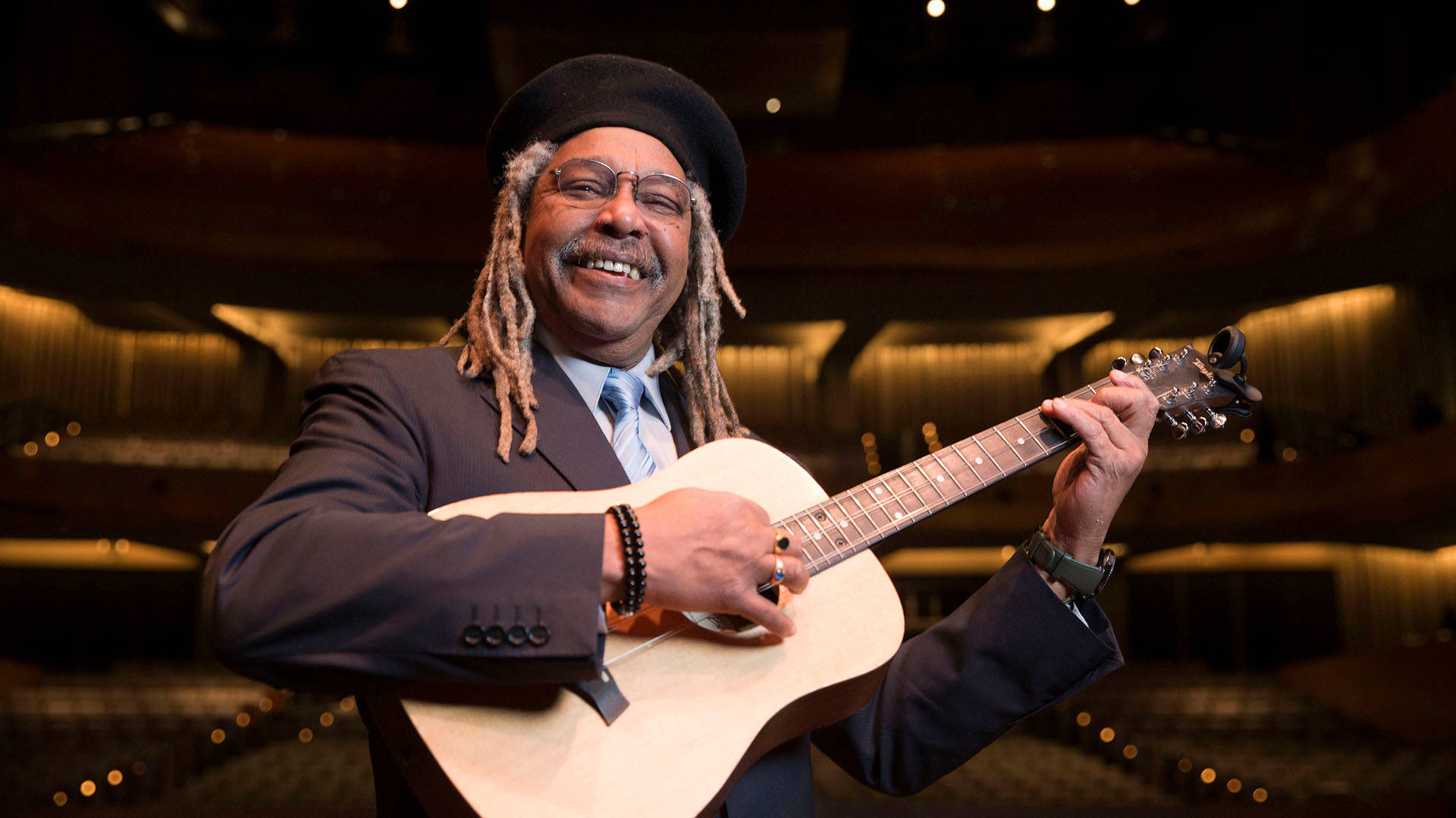 A man smiling for the camera and holding a guitar. He has dreadlocks, a black hat, and a suit on. 