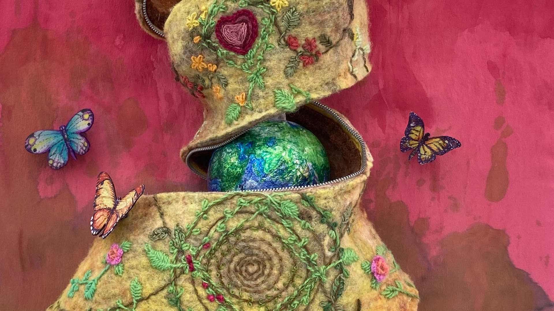 Close up of a felted art piece by Carolyn Benforado. Three butterflies against a pink red background. In the center, an Earth globe is being "unzipped" from a brown and green fabric.