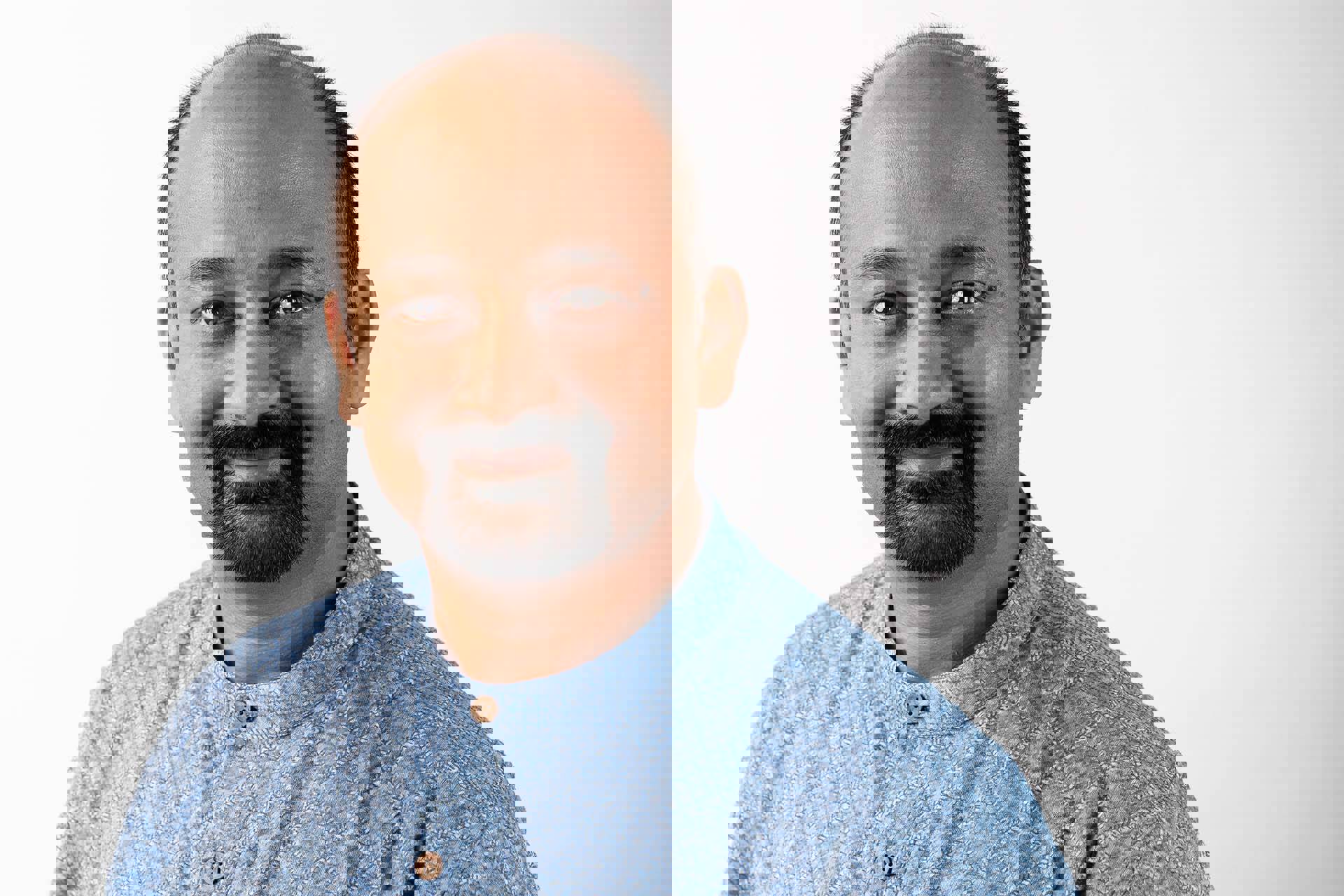 Headshot of a man with medium complexion, a bald head and dark hair and goatee. He is wearing a blue button up shirt.