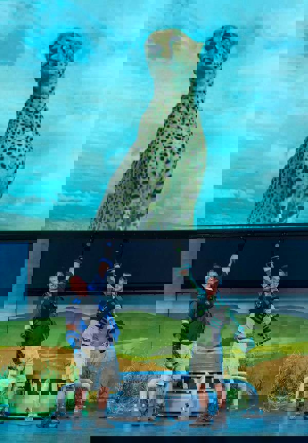 Kratt brothers, two light complexion men wearing shorts, hiking boots, and superhero vests/gloves, point to a cheetah image on a slideshow screen during a LIVE 2.0 performance.